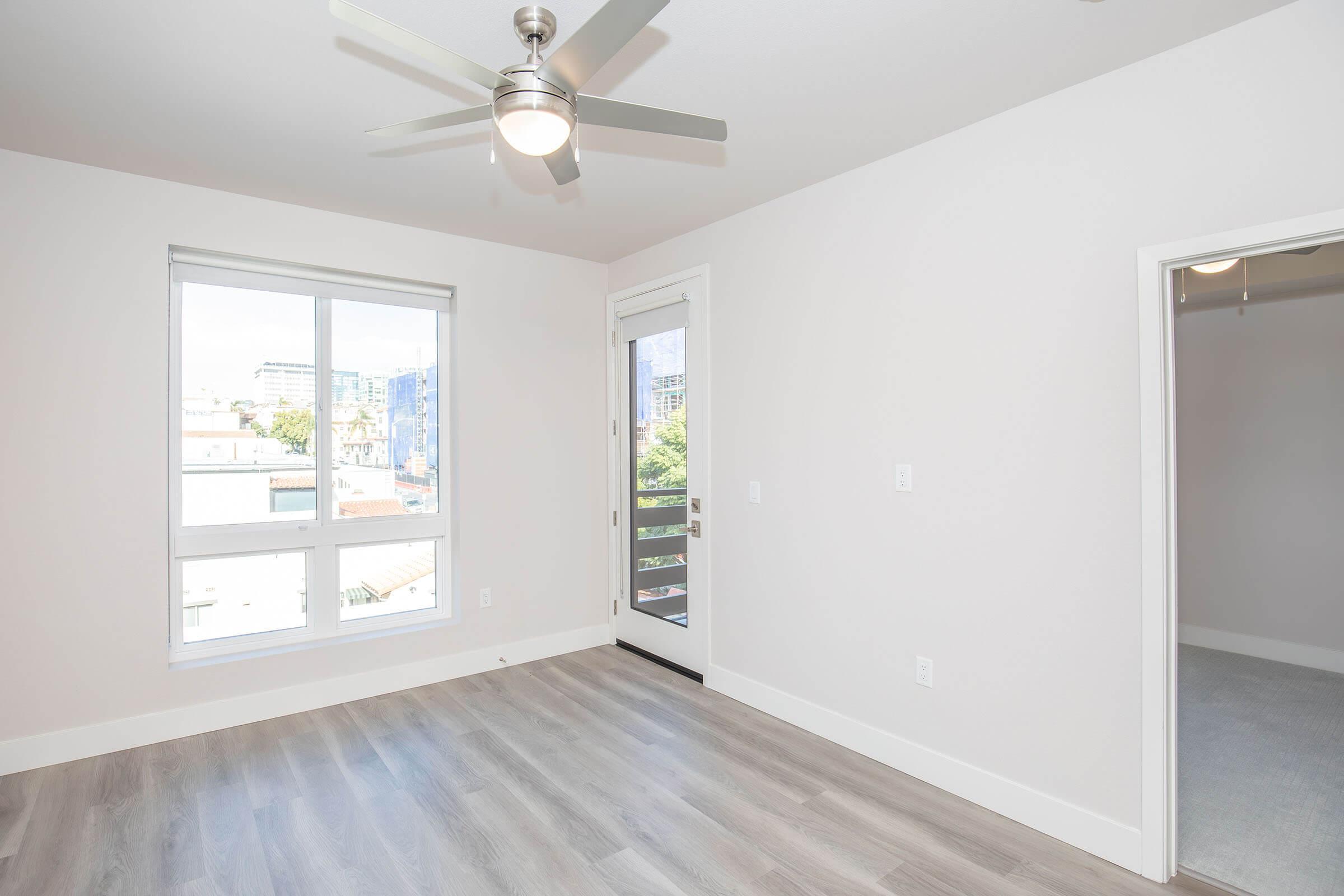 Bright and airy room featuring light-colored walls, a ceiling fan, and large windows providing natural light. A door leads to a balcony, and there’s a doorway on the right side, possibly leading to a closet or another room. The floor is light wood laminate, creating a modern and inviting atmosphere.