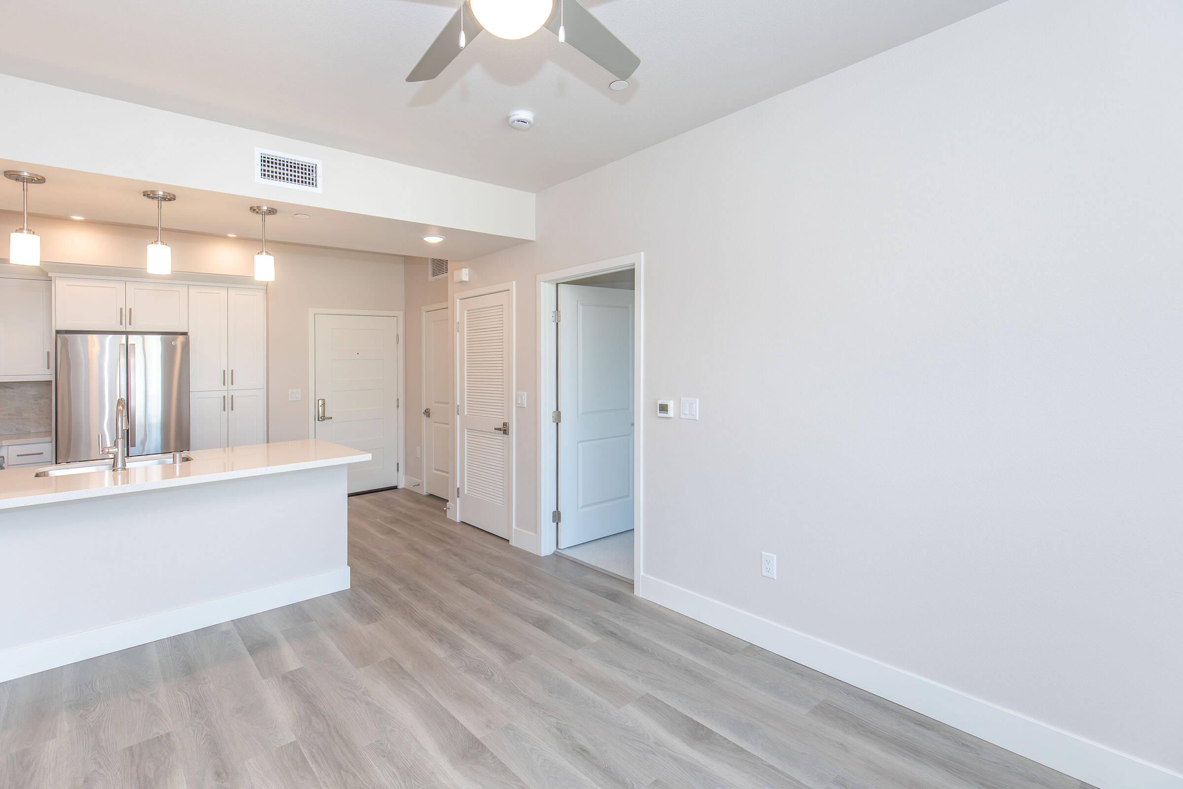 A modern, minimalist apartment interior featuring light-colored walls, hardwood flooring, and an open layout. The kitchen area has white cabinets, stainless steel appliances, and pendant lighting. A doorway leads to a second room, and there is a ceiling fan overhead, adding a contemporary touch to the space.