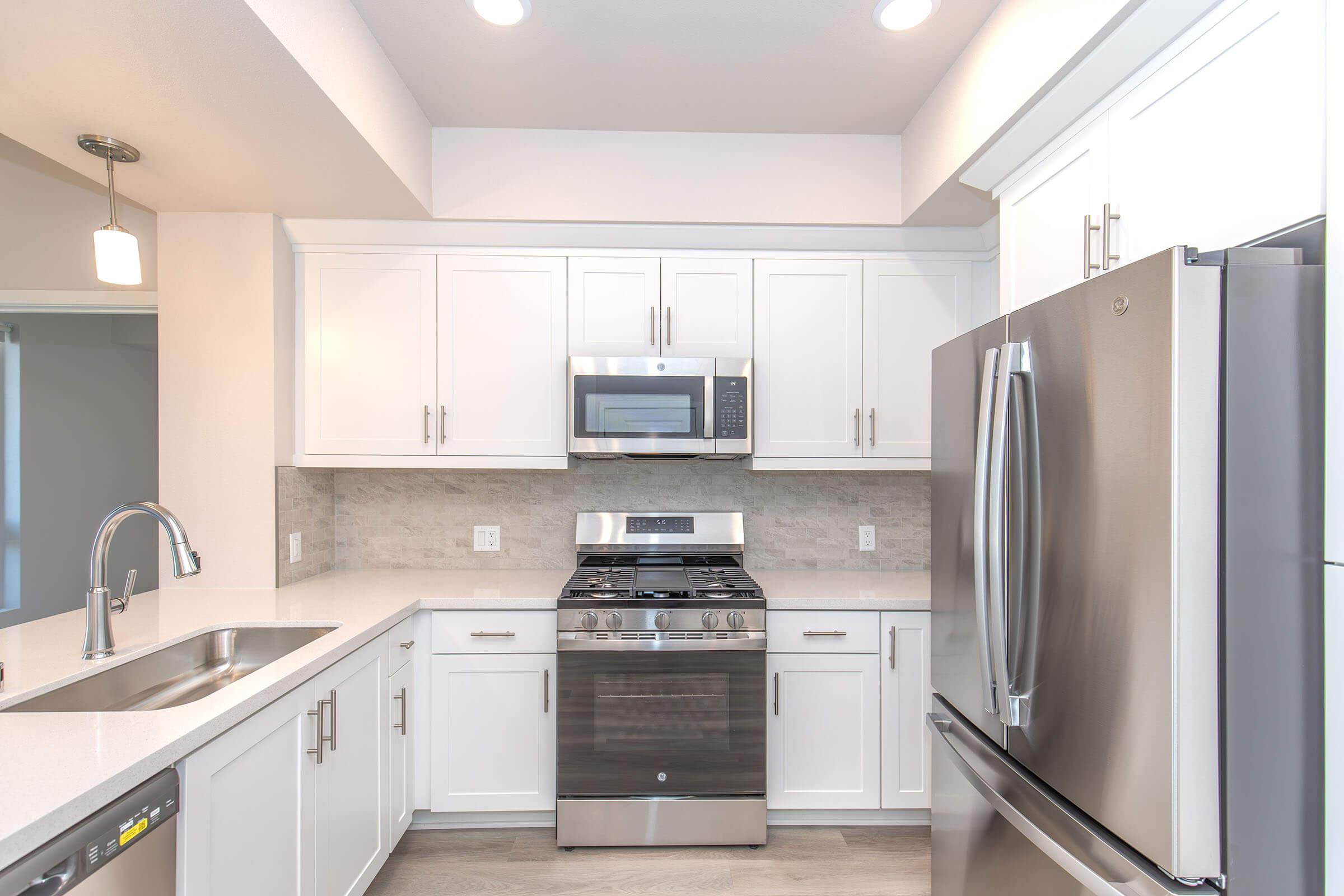 Modern kitchen featuring white cabinetry, stainless steel appliances including a refrigerator and gas stove, a microwave above the stove, and a single-basin sink. The countertop is a light, solid surface, and the backsplash is a neutral stone design. Natural light fills the space.