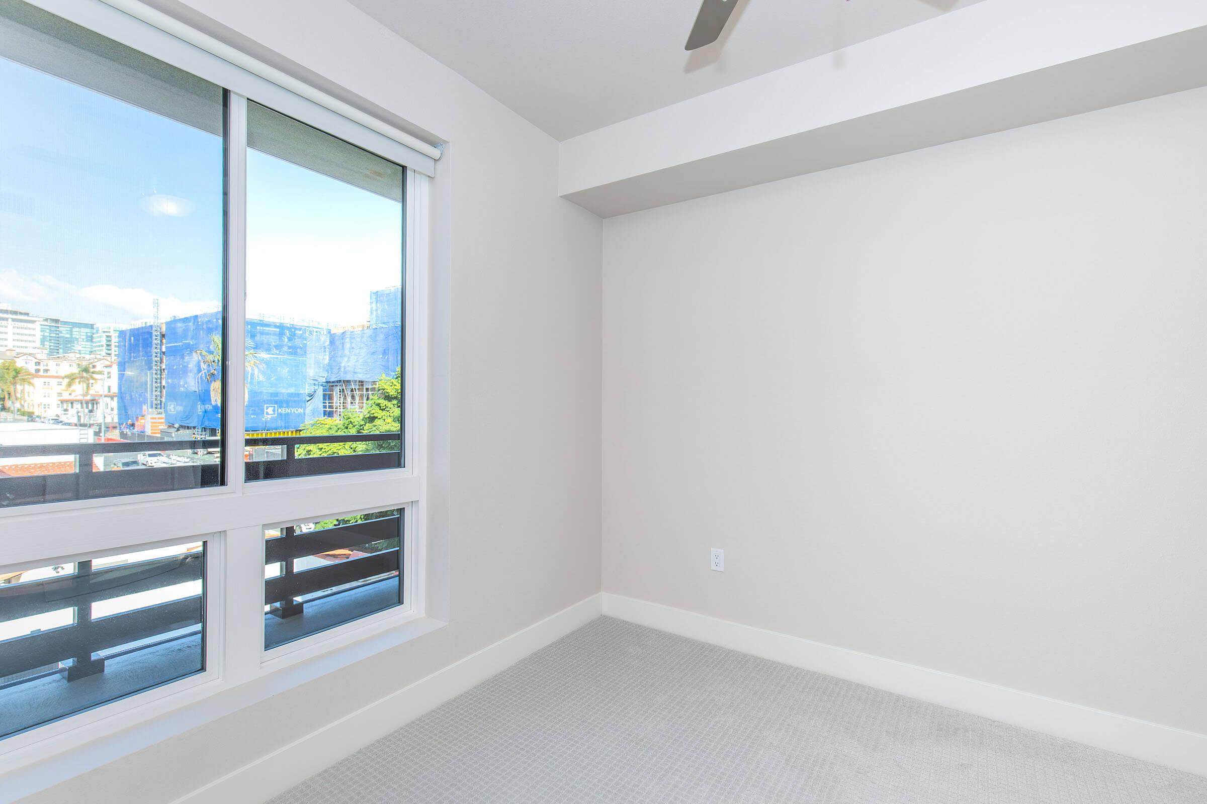A bright, empty room with a large window showcasing a view of a cityscape. The walls are painted a light color, and there is a ceiling fan visible. The floor is covered with light-colored carpet, creating a clean and modern aesthetic.