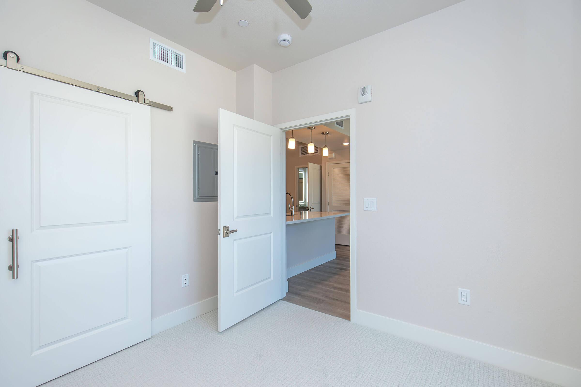 Interior view of a modern room featuring an open door leading to another space. The walls are painted light colors, and there's a ceiling fan visible. The floor has light-colored planks, and a doorway in the background suggests connectivity to another area, likely a kitchen or bathroom.