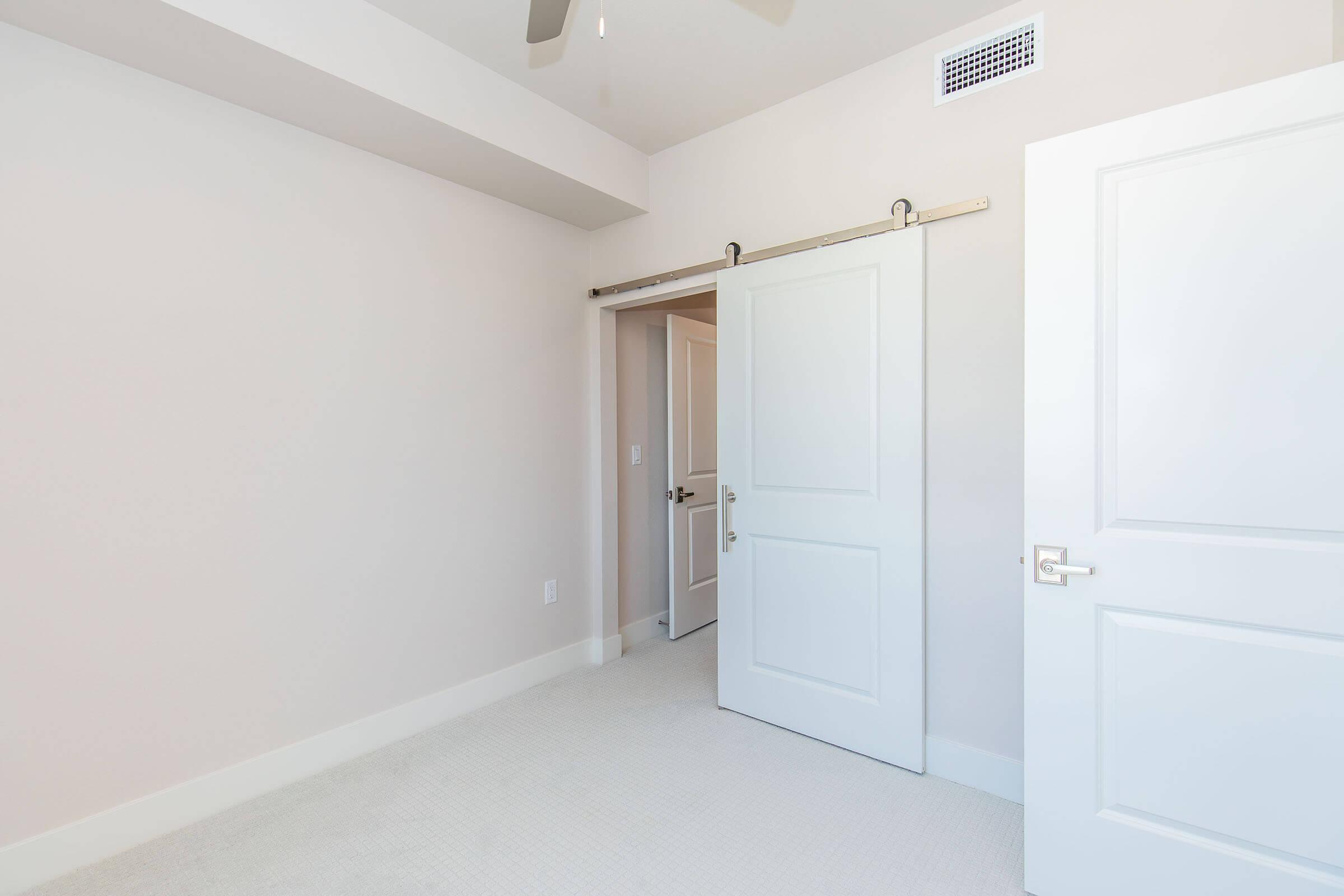 A bright, minimalistic interior of a room featuring a double door with a sliding barn door track, a ceiling fan, and neutral-colored walls. The flooring is carpeted, and there is an open doorway leading to another room. Natural light fills the space, creating a clean and inviting atmosphere.