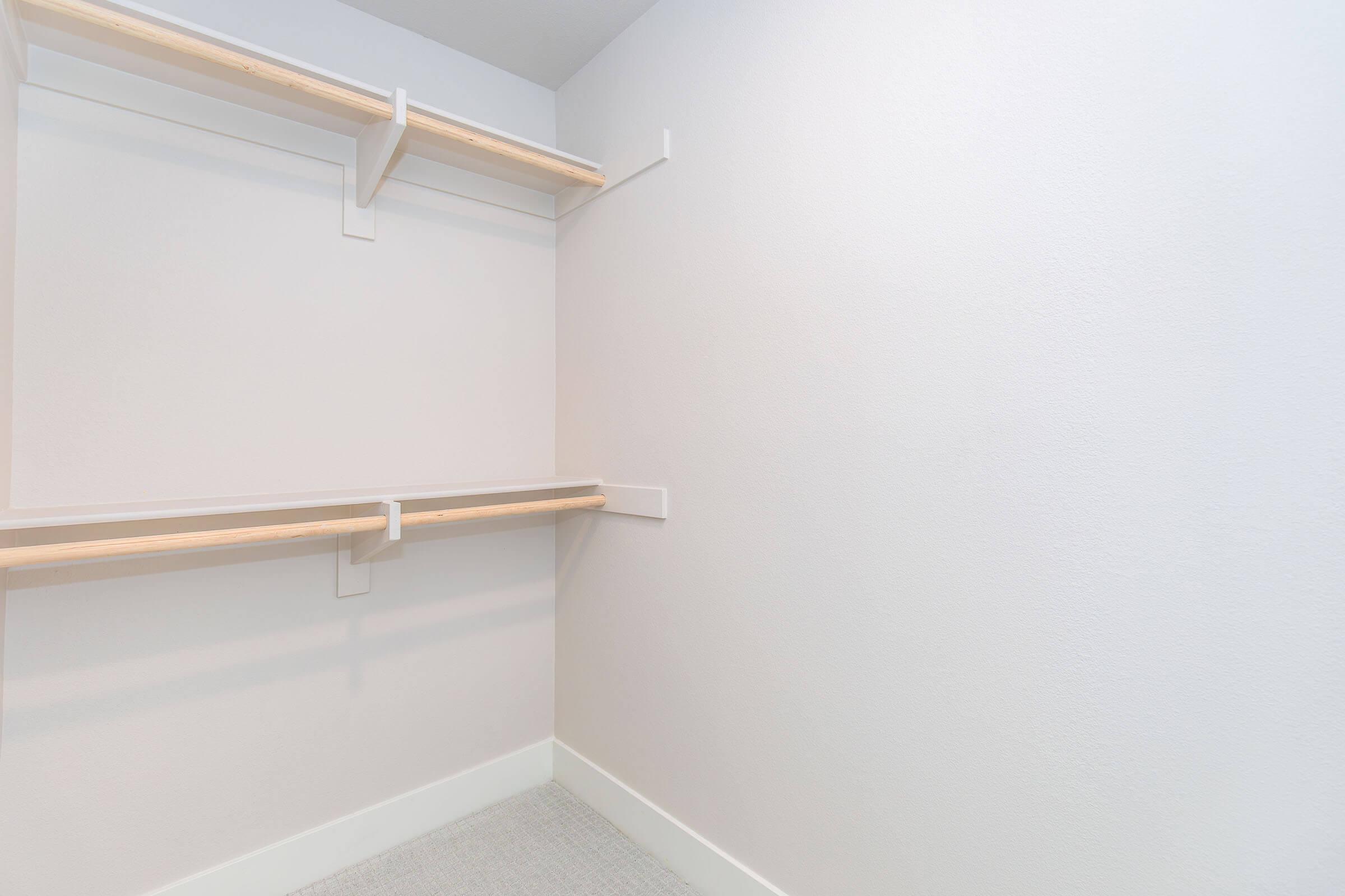 An empty walk-in closet featuring light gray walls and two wooden shelves mounted on the side. The floor is covered with carpet, and there is ample space for storage. The overall look is clean and minimalist.
