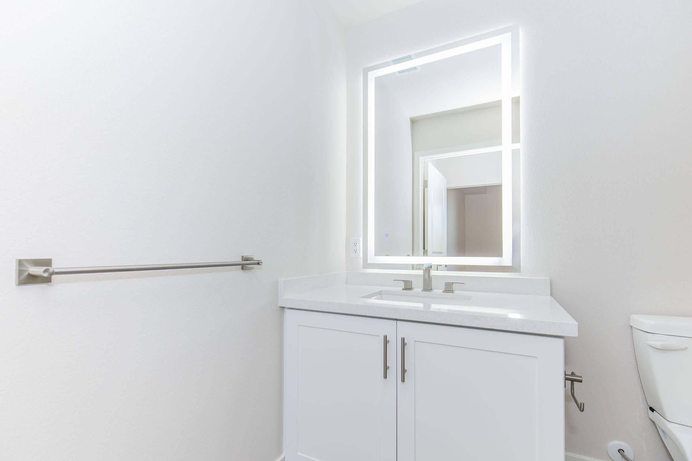 Modern bathroom featuring a sleek double sink vanity with a white countertop, illuminated mirror, and towel bar. A clean, minimalist design with white walls and a toilet in the corner, highlighting the spacious and bright atmosphere of the room.
