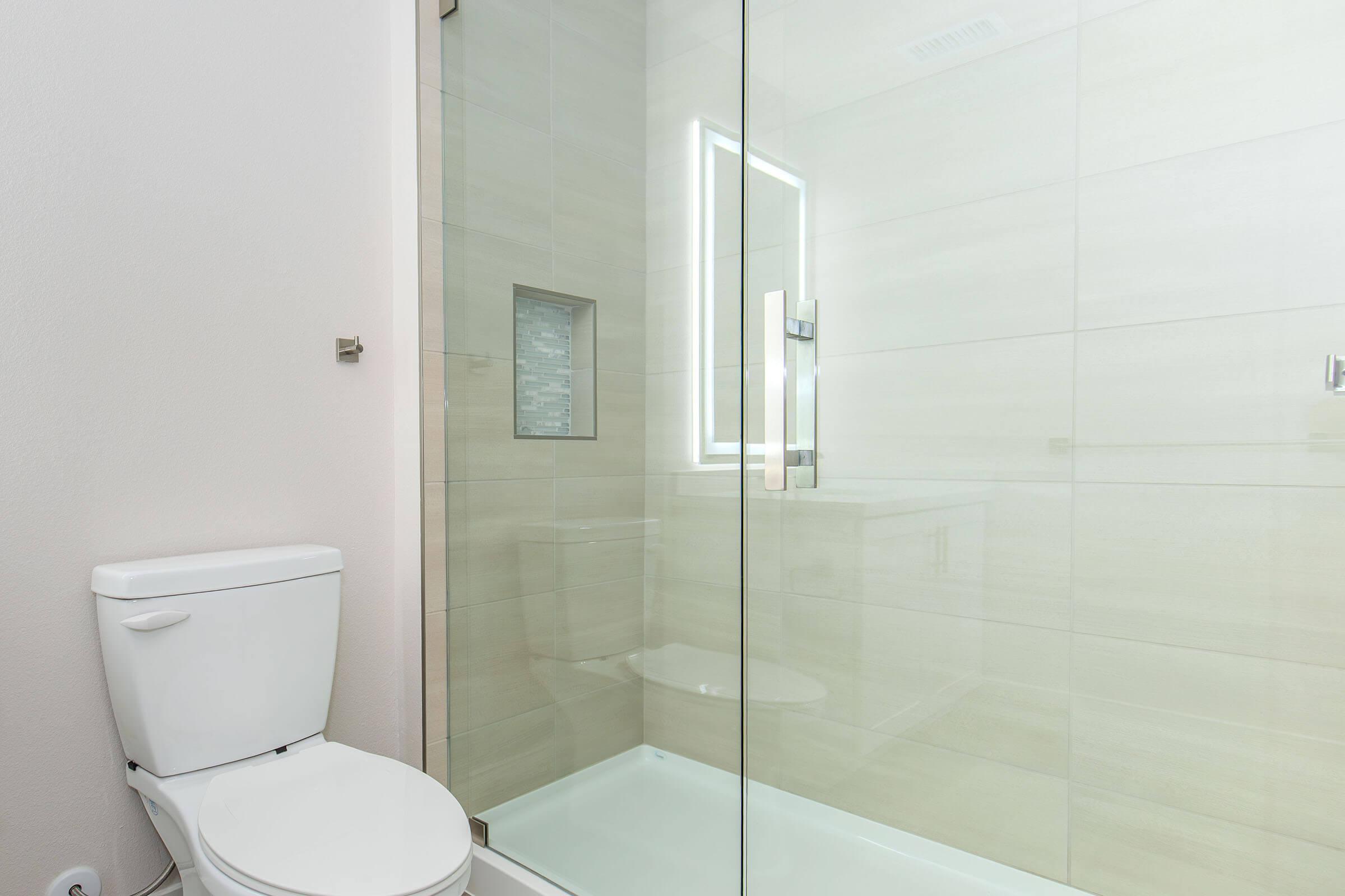 Modern bathroom featuring a glass shower enclosure and a white toilet. Neutral-toned tiles cover the walls and floor. A small window is present in the shower area, providing natural light. The overall design is sleek and minimalistic, emphasizing cleanliness and simplicity.