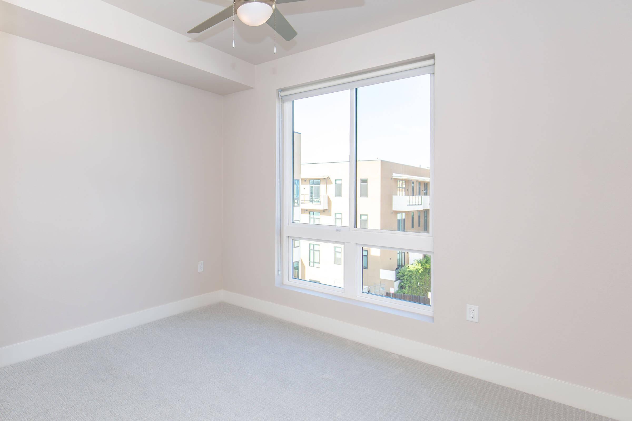 A bright, empty room with light-colored walls and a ceiling fan. A large window offers a view of neighboring buildings, allowing natural light to enter. The floor is carpeted, and there are no furnishings, creating a spacious and airy feel.