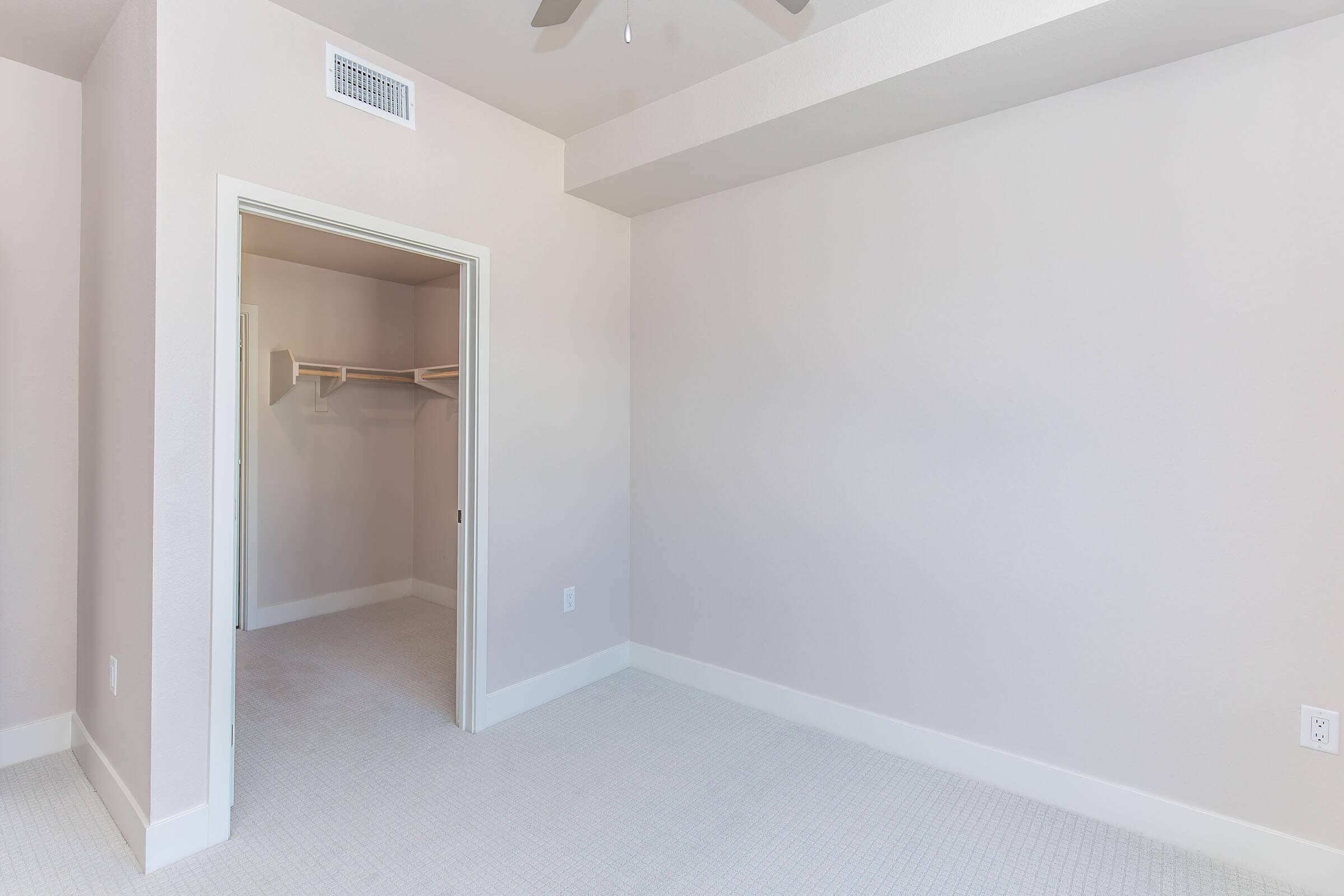 Empty room with light beige walls and a ceiling fan. A doorway on the left leads to a small closet with shelves. The floor is covered in soft, light-colored carpeting, creating a bright and spacious feel.
