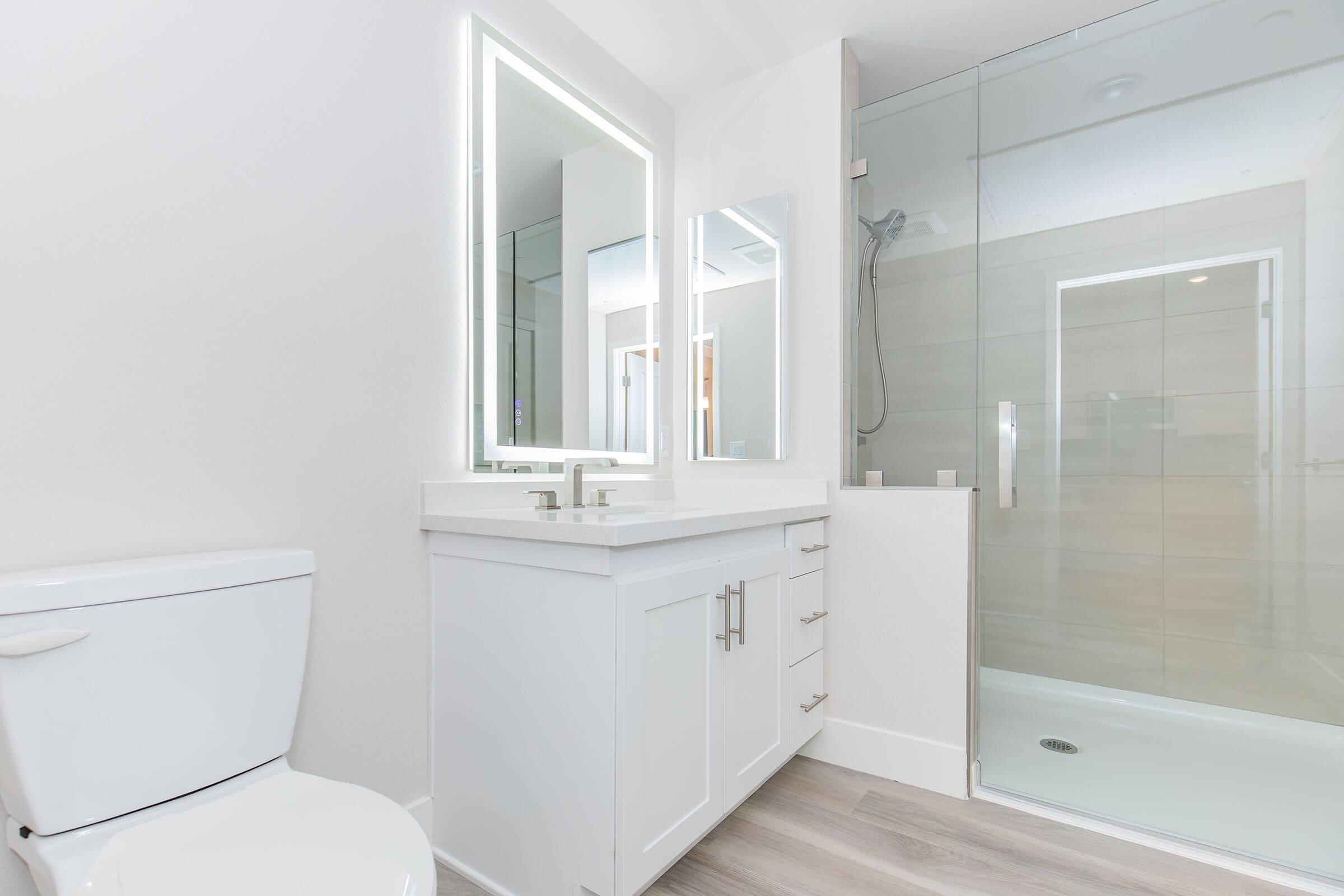 Modern bathroom featuring a white toilet, a sleek vanity with a large mirror and backlighting, and a glass shower enclosure. The decor is minimalistic with light-colored walls and wood-like flooring, creating a clean and spacious atmosphere.