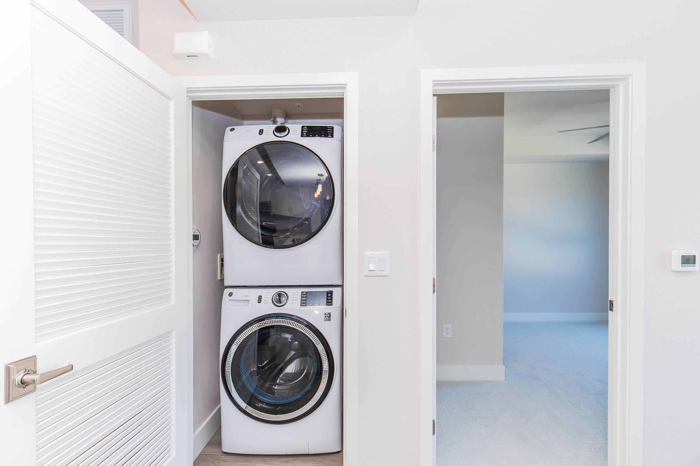 A laundry area featuring a stacked washer and dryer unit in a bright, modern space. The entrance has white doors, and adjacent is an empty room with light-colored walls and carpet, showcasing a minimalistic and clean aesthetic.