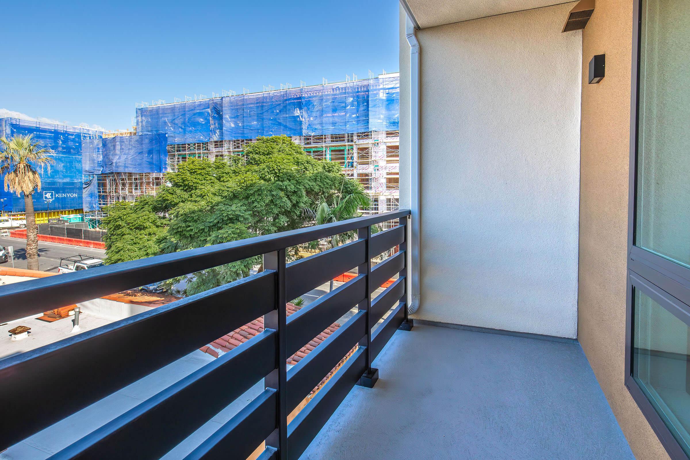 A modern balcony with a black railing overlooking a construction site covered in blue tarps. In the foreground, a tree is visible, and the bright blue sky creates a vibrant backdrop. The scene suggests an urban environment with ongoing development activities.