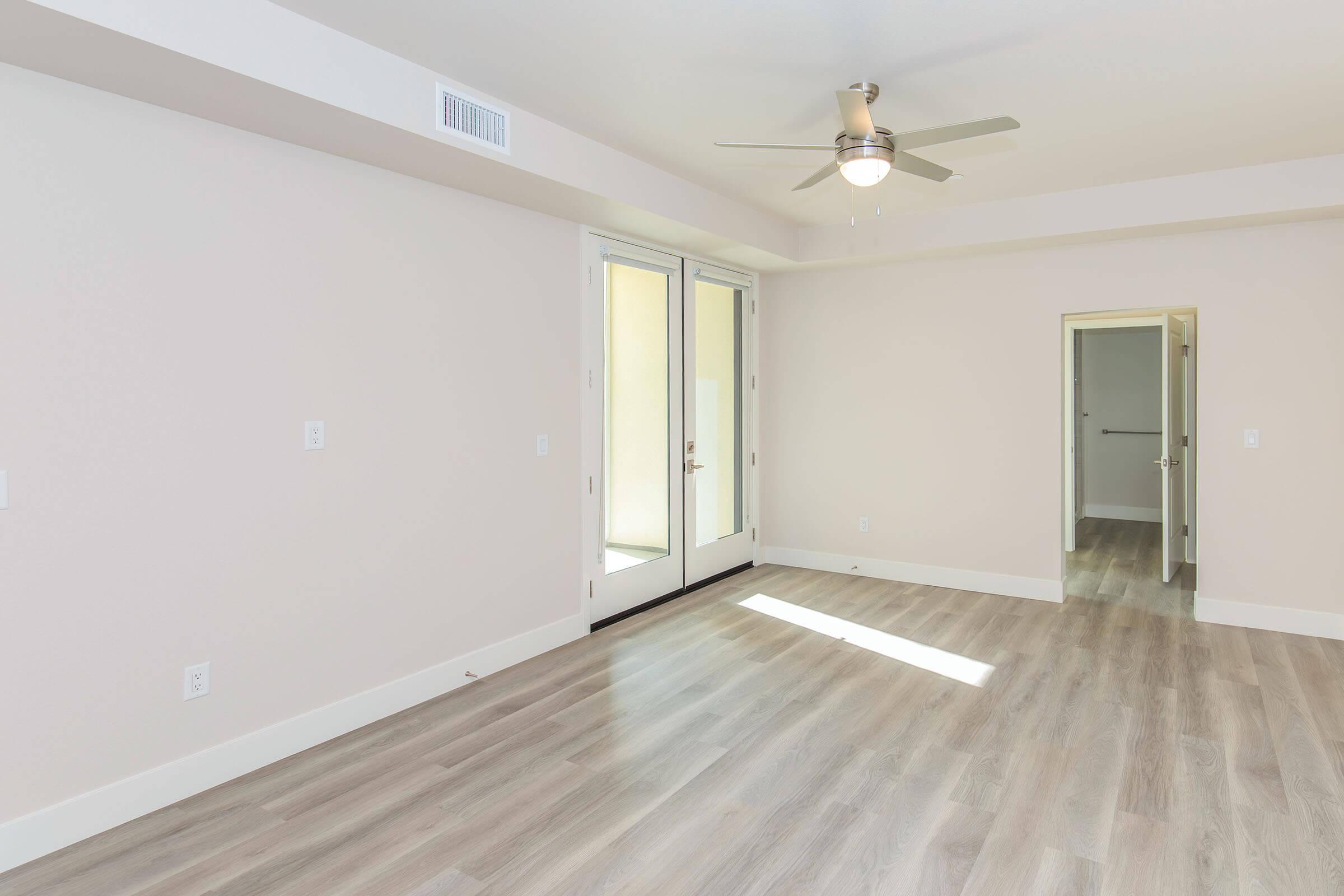 A spacious, empty room with light-colored walls and hardwood flooring. It features a ceiling fan and a set of double doors leading to a balcony. There is a doorway on the right side, possibly leading to a closet or another room. Natural light brightens the space.