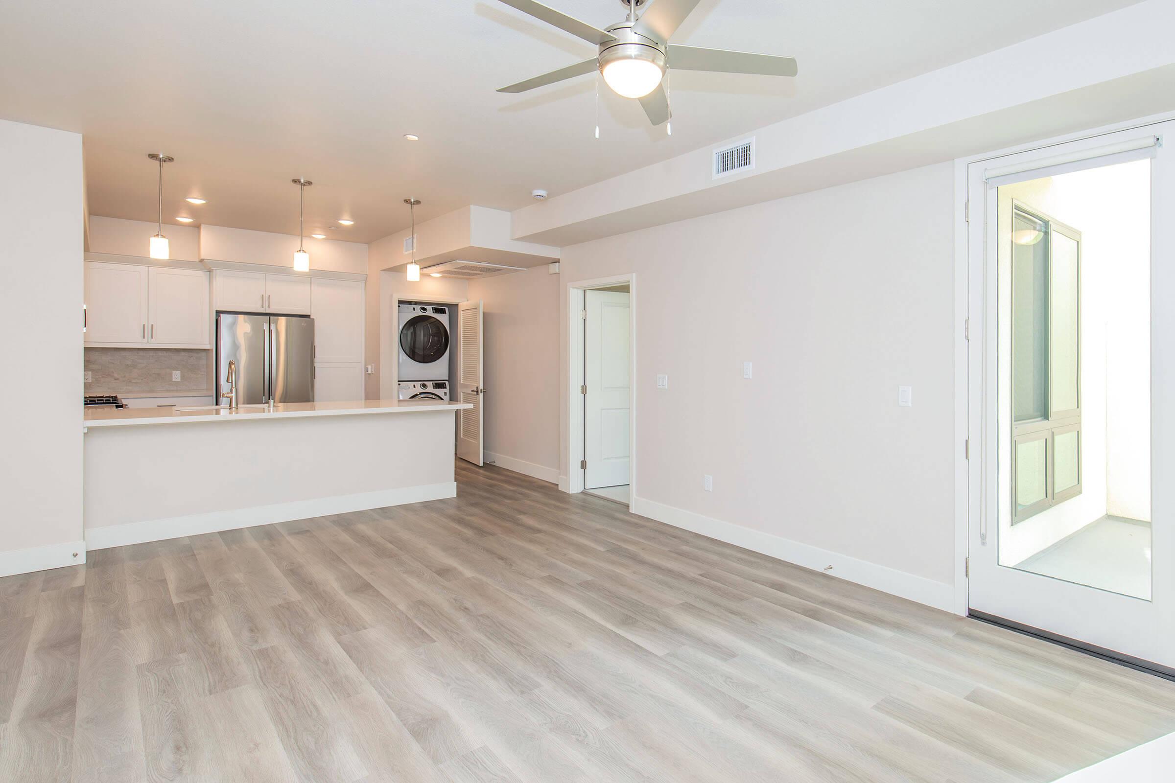 Modern open-concept living space featuring light wood flooring, white walls, and a ceiling fan. The kitchen is equipped with stainless steel appliances and a breakfast bar. A door leads to an outdoor area, providing natural light and a view.