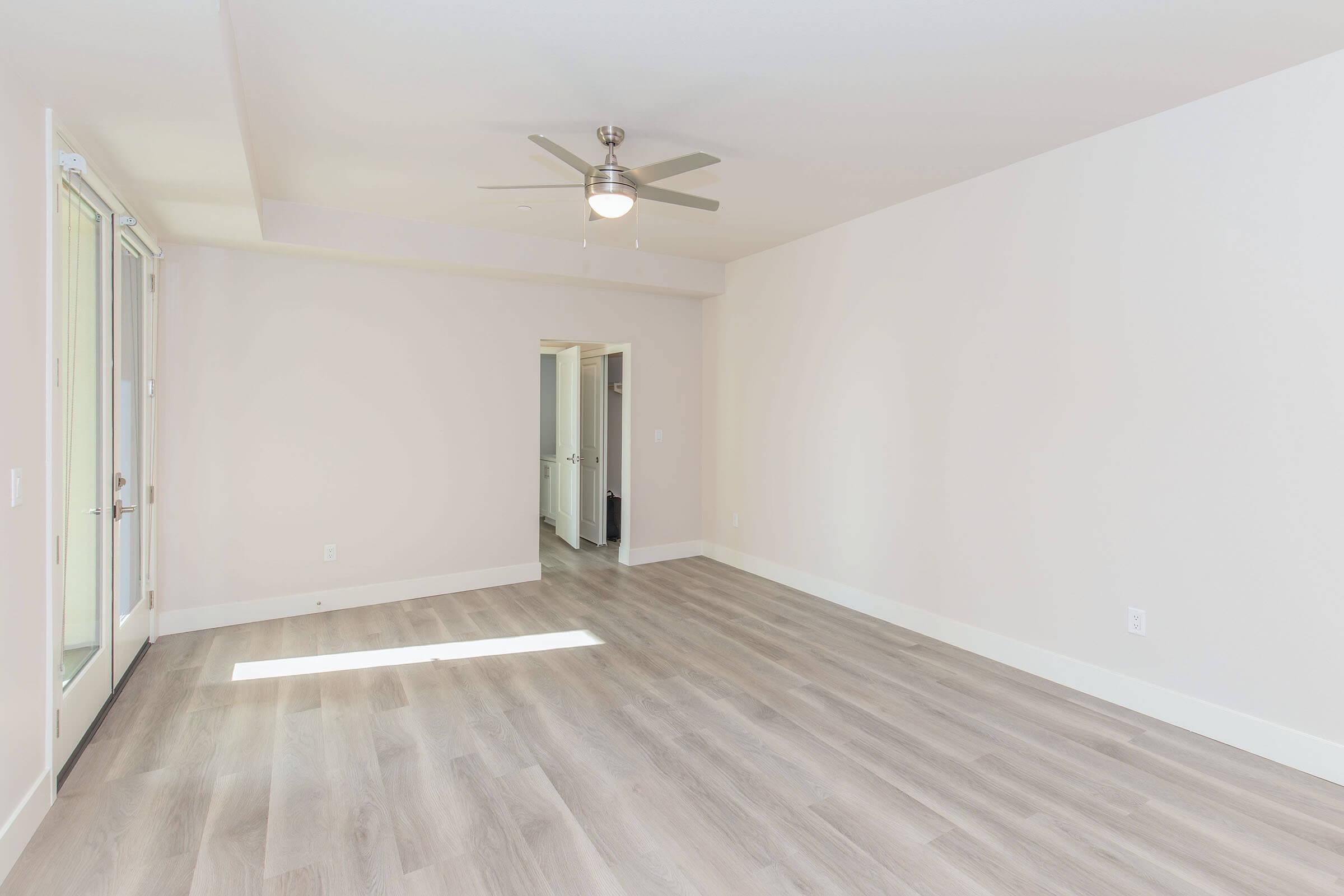 A spacious, empty room with light-colored walls and a light wooden floor. A ceiling fan is installed, and there are large windows allowing natural light to enter. A doorway leads to another room, creating an open and airy atmosphere.