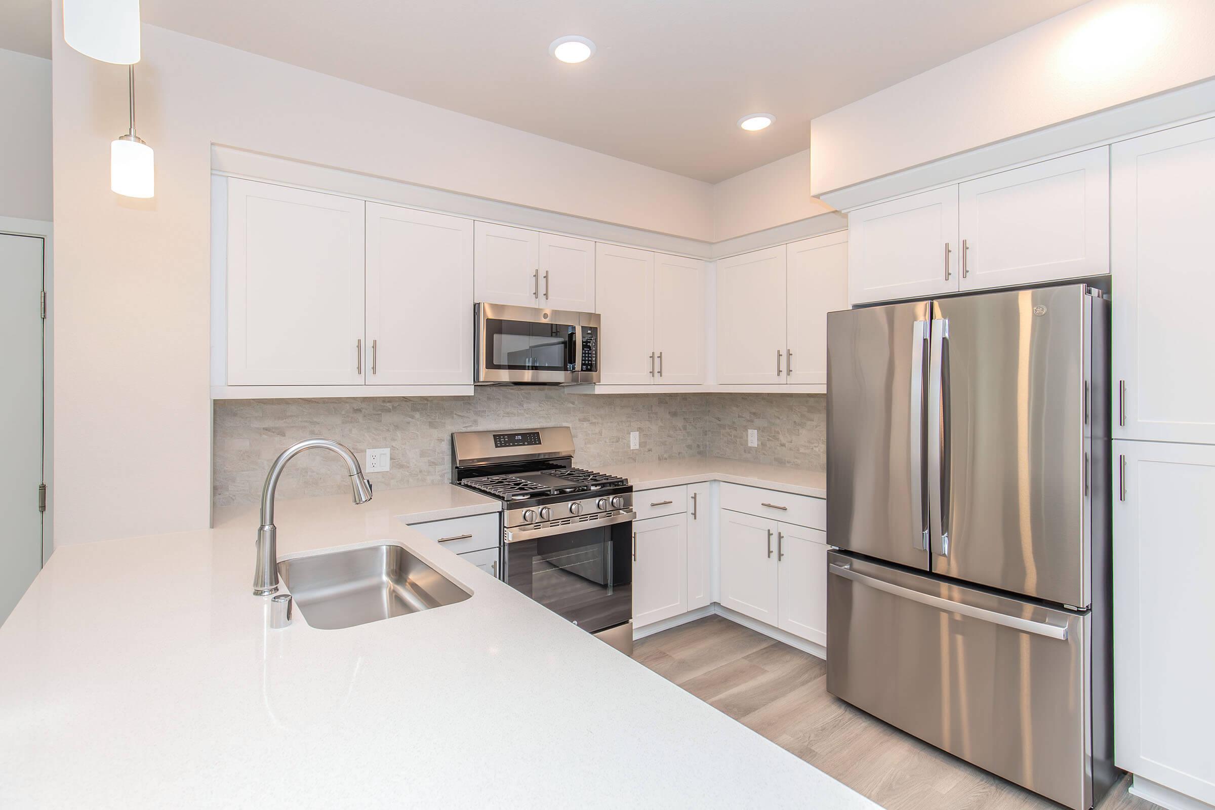 Modern kitchen featuring white cabinets, stainless steel appliances including a refrigerator and microwave, a gas stove, and a sleek sink. The countertop is light-colored with a smooth finish, and the space is well-lit with recessed lighting and pendant lights. The flooring is light wood laminate.