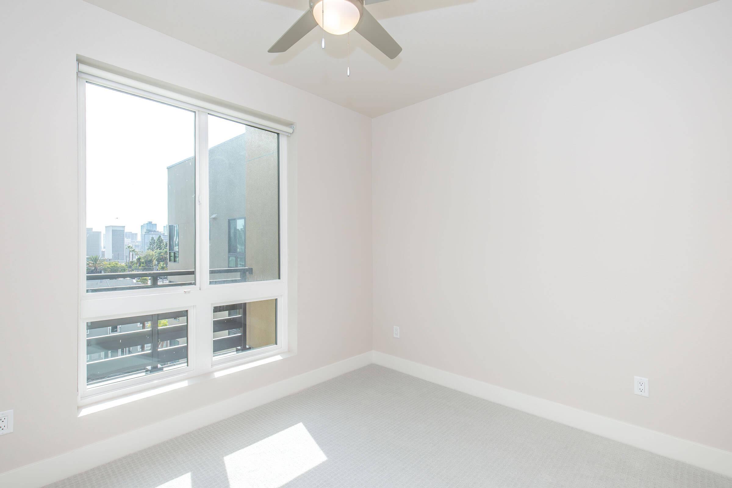 Bright, empty bedroom featuring light walls and a ceiling fan. A large window offers a view of the city skyline. The floor is covered with neutral-colored carpet, creating a clean and spacious atmosphere.