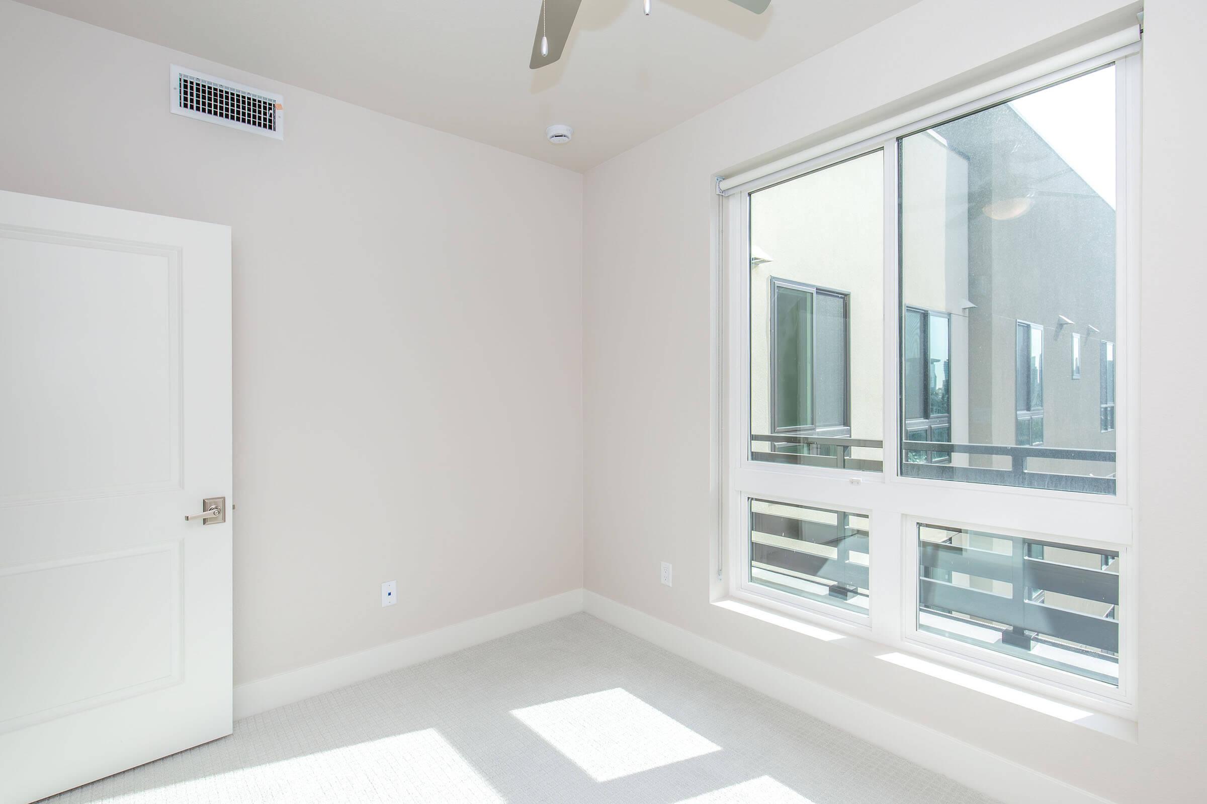 A well-lit corner of a minimalistic room featuring a large window with modern outdoor views. The wall is painted in a soft, neutral color, and a ceiling fan is visible above. The space has a clean carpeted floor and a simple white door leading to another area.