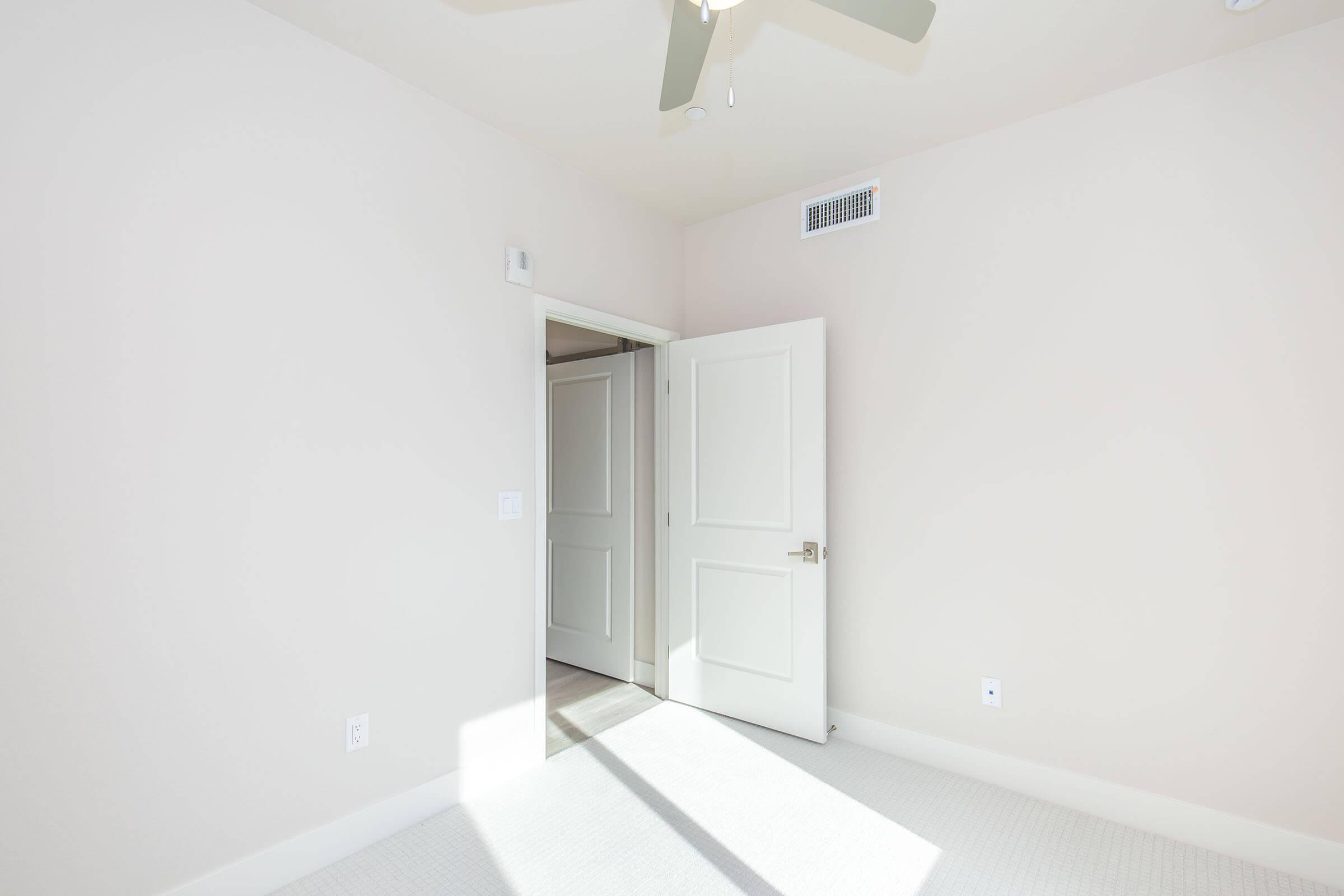 Bright and airy room with light-colored walls and a ceiling fan. A white door is partially open, leading to another room. The floor is light-colored, and the space is minimally furnished, creating a clean and inviting atmosphere.