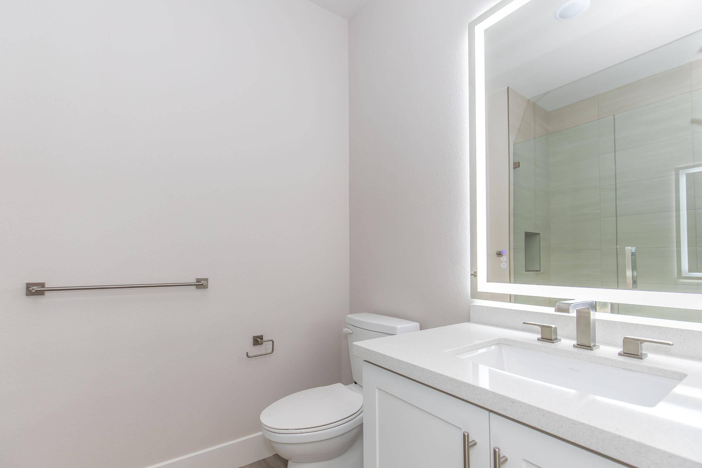 A modern bathroom featuring a white toilet, a sleek vanity with a stone countertop and two faucets, a mirror with backlighting, and a glass walk-in shower. The walls are painted in a light color, and there's a towel bar mounted on the wall. The overall design is clean and minimalist.