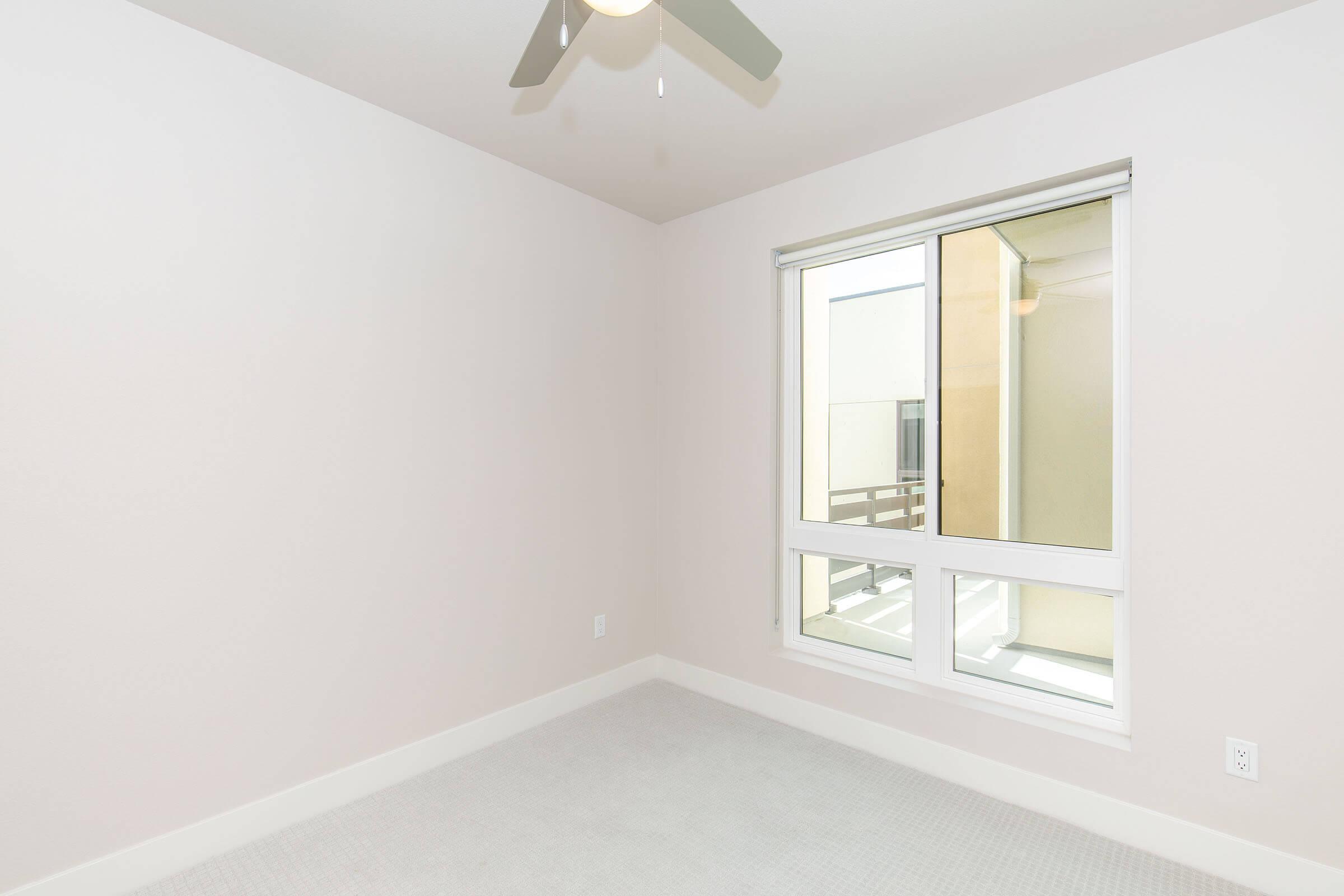 A blank bedroom with light-colored walls and a ceiling fan. There is a large window allowing natural light, with a view of a balcony outside. The carpeted floor is neutral in color, creating a spacious and minimalist atmosphere.