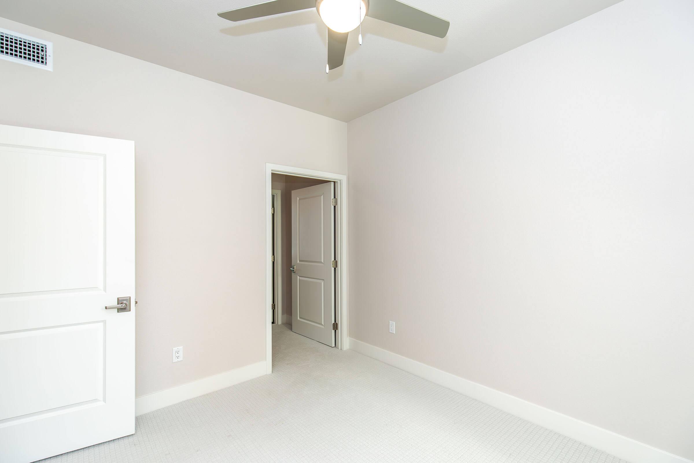 A simple, empty room featuring light-colored walls, a ceiling fan, and a light-colored carpet. There are two doors—one leading to an adjacent room and the other presumably leading outside. The room appears well-lit and minimalistic, suitable for various uses.