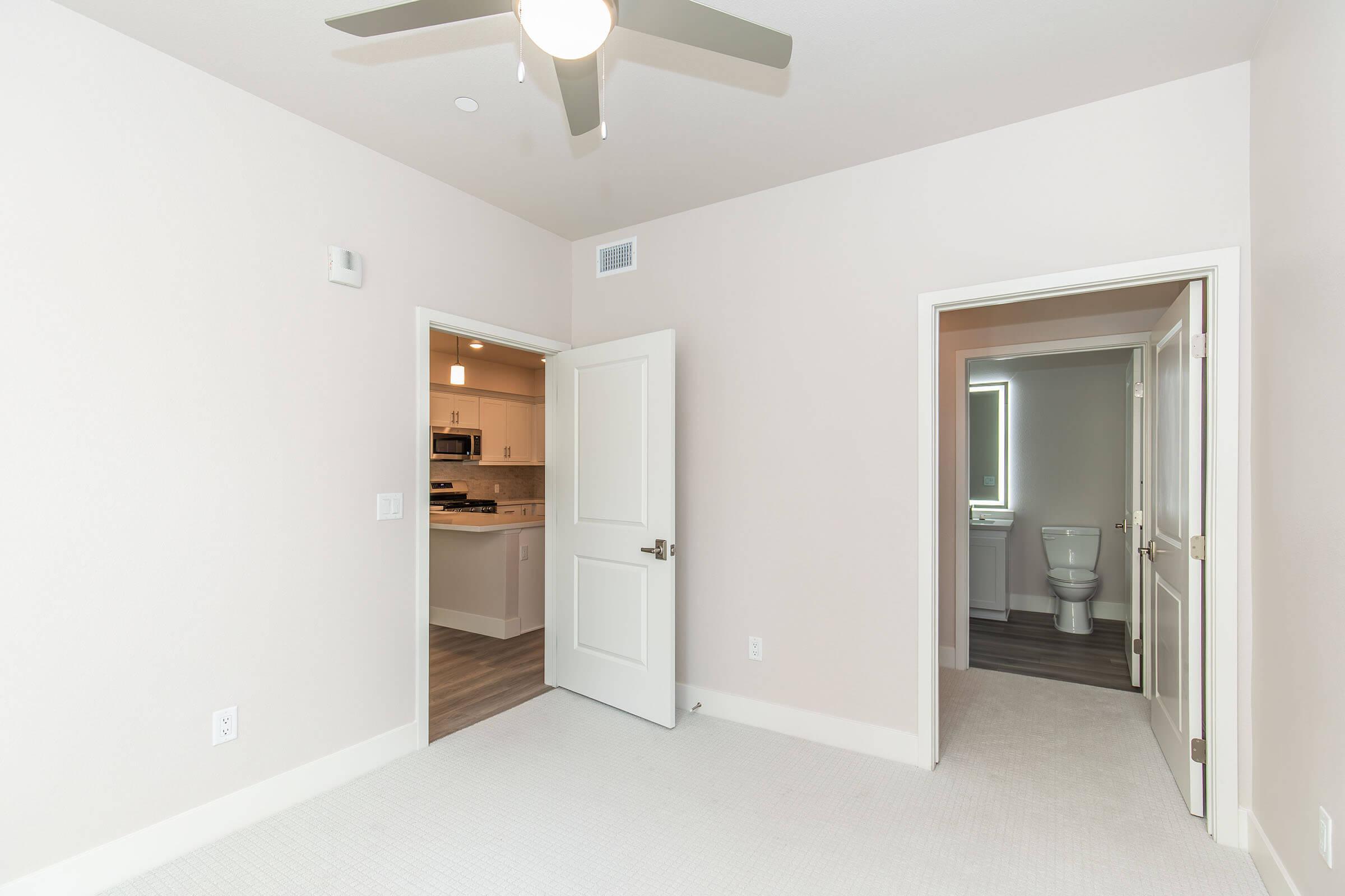 A light-colored, empty room with beige walls and carpet. A ceiling fan is mounted above, and a doorway on the left leads to a kitchen area with visible appliances. Another doorway on the right opens into a bathroom, which includes a toilet and sink. The overall space is well-lit and modern.