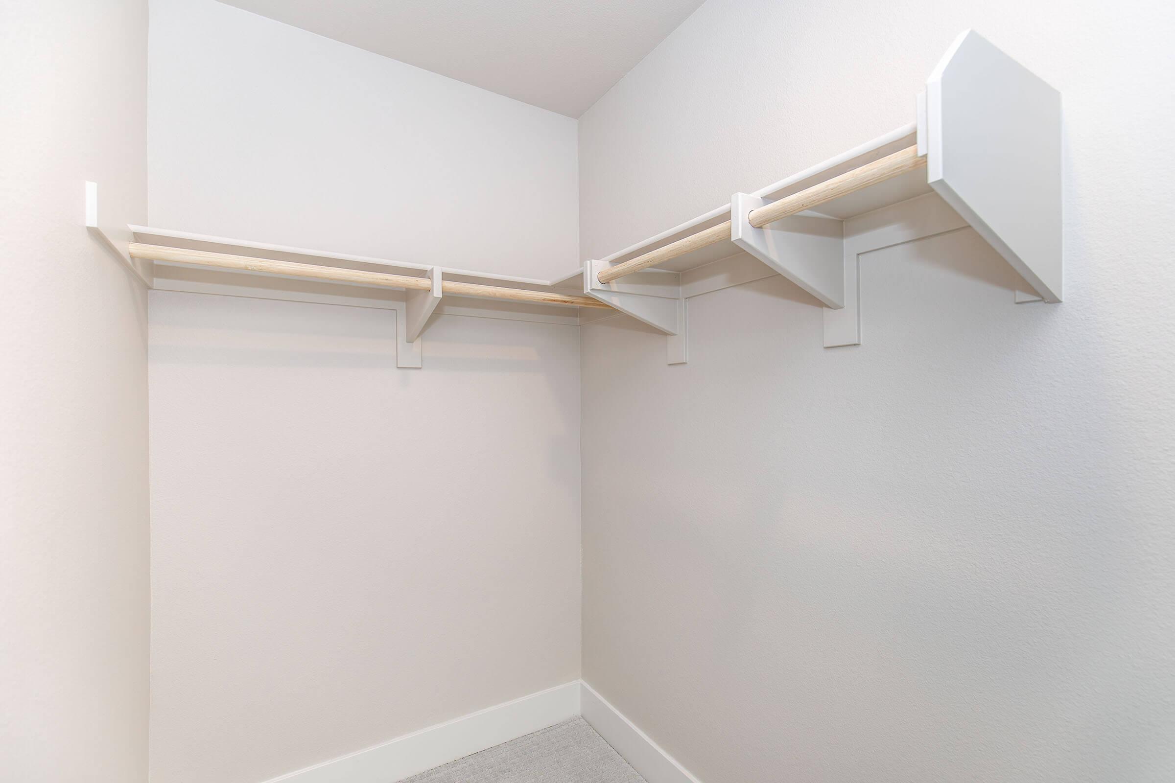 Empty corner closet with white walls and light wood shelving. The closet features three shelves mounted on the wall and space for hanging clothes, creating a clean and minimalist storage area.