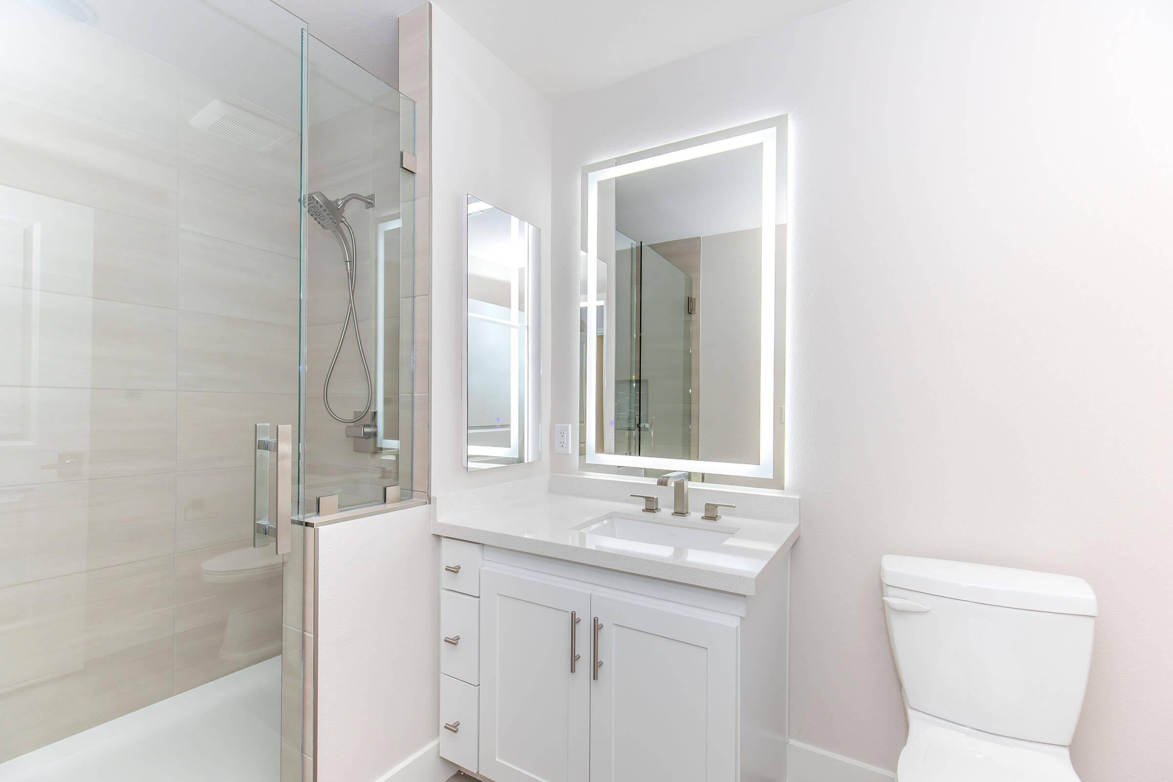 Modern bathroom featuring a glass shower, a sleek double sink vanity with silver fixtures, illuminated mirror, and a white toilet. The walls are light-colored, creating a spacious and clean appearance. Natural light enhances the bright atmosphere.