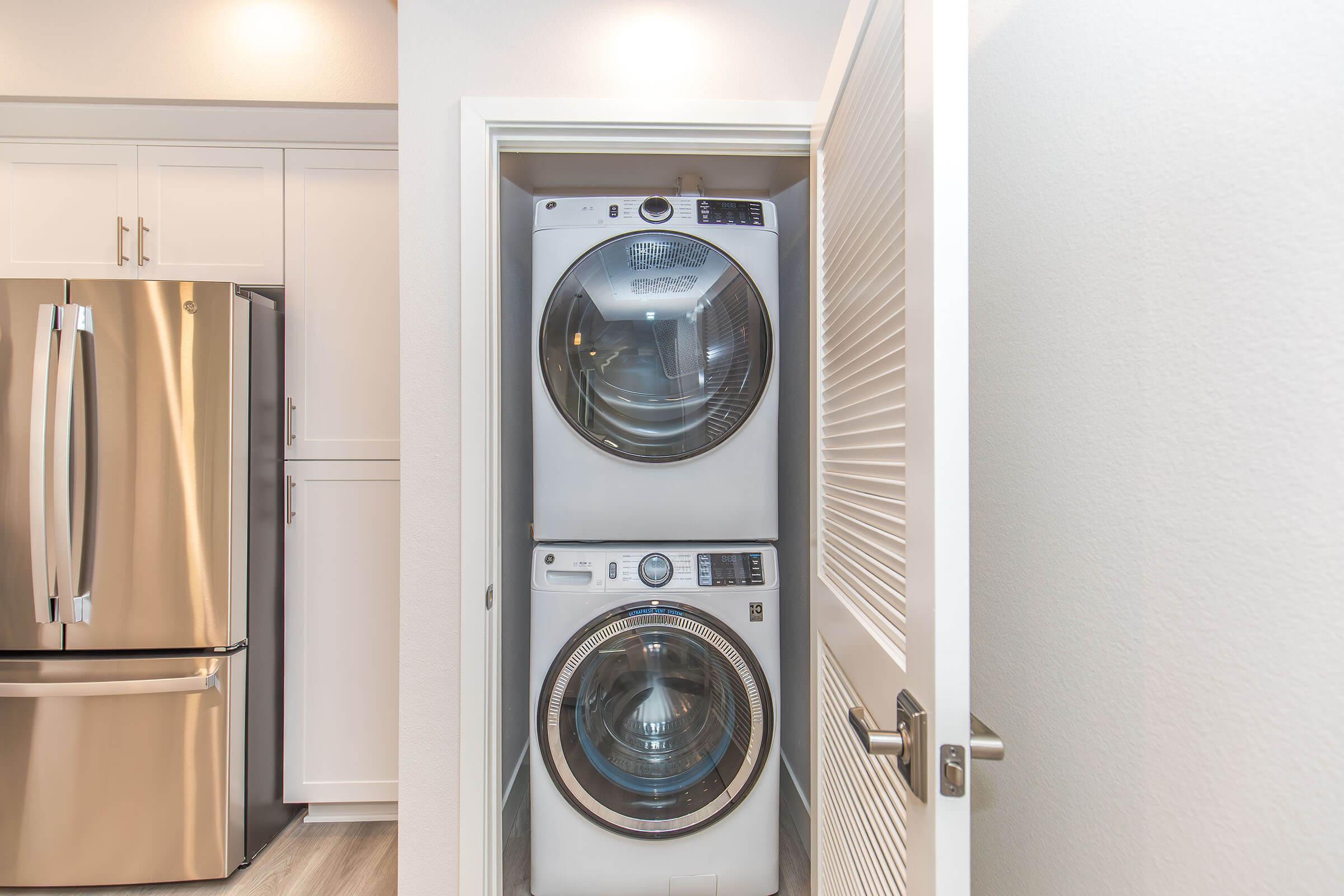 A stacked washer and dryer unit in a kitchen or laundry area, with modern stainless steel appliances, including a refrigerator visible nearby. The space features light-colored walls and flooring, enhancing the bright and clean look of the room.