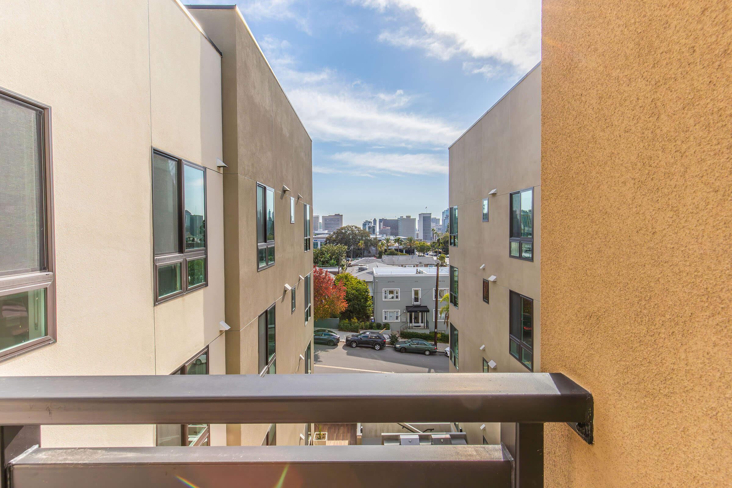 View from a balcony between two modern buildings, showcasing a glimpse of a city skyline in the distance. The foreground includes angular building designs, while the background features trees and a clear blue sky. The overall scene captures a blend of urban architecture and natural elements.