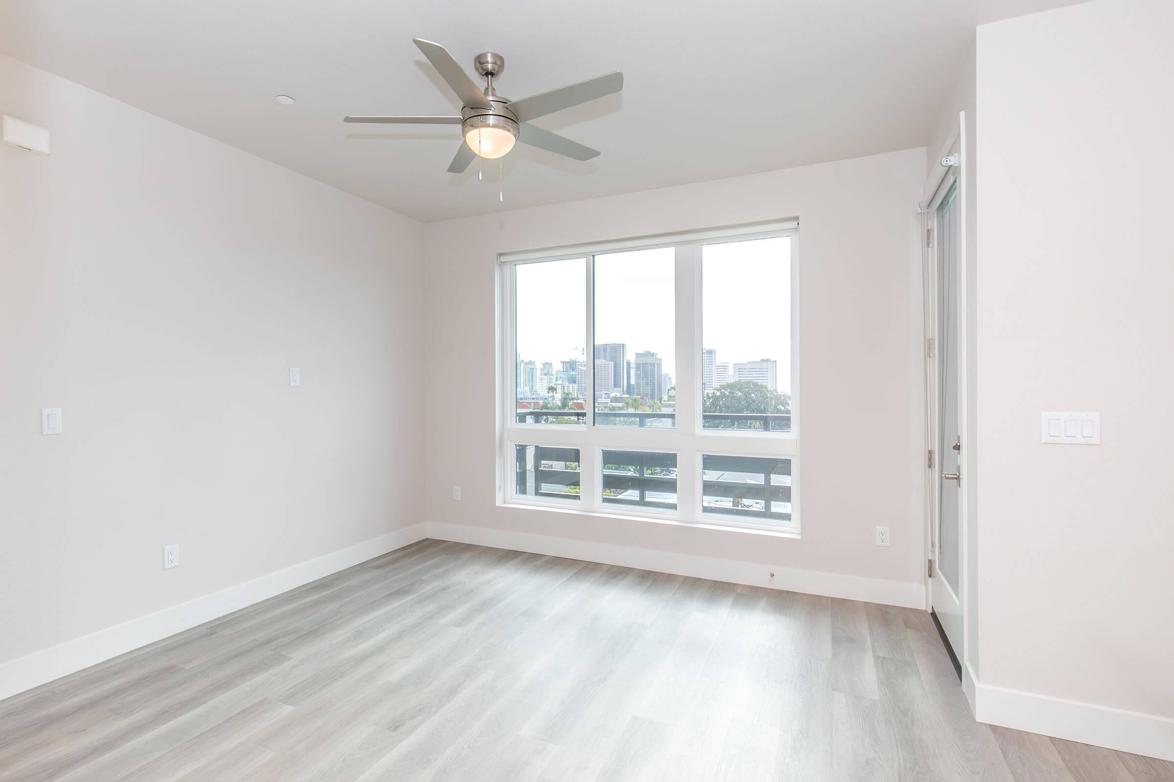 A bright, empty room featuring light-colored walls and hardwood flooring. A ceiling fan is visible, and there is a large window showcasing a view of a cityscape. The room has a modern design with minimal decor, making it ideal for personal customization.