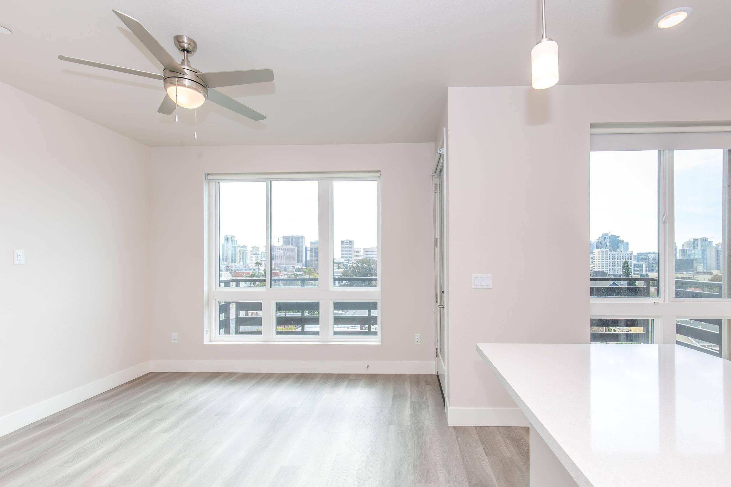 Bright, modern interior of an apartment featuring large windows with city views, light-colored walls, a ceiling fan, and a sleek kitchen island. Soft natural light floods the space, highlighting the contemporary design and flooring. Walls are unadorned, providing a fresh and open atmosphere.