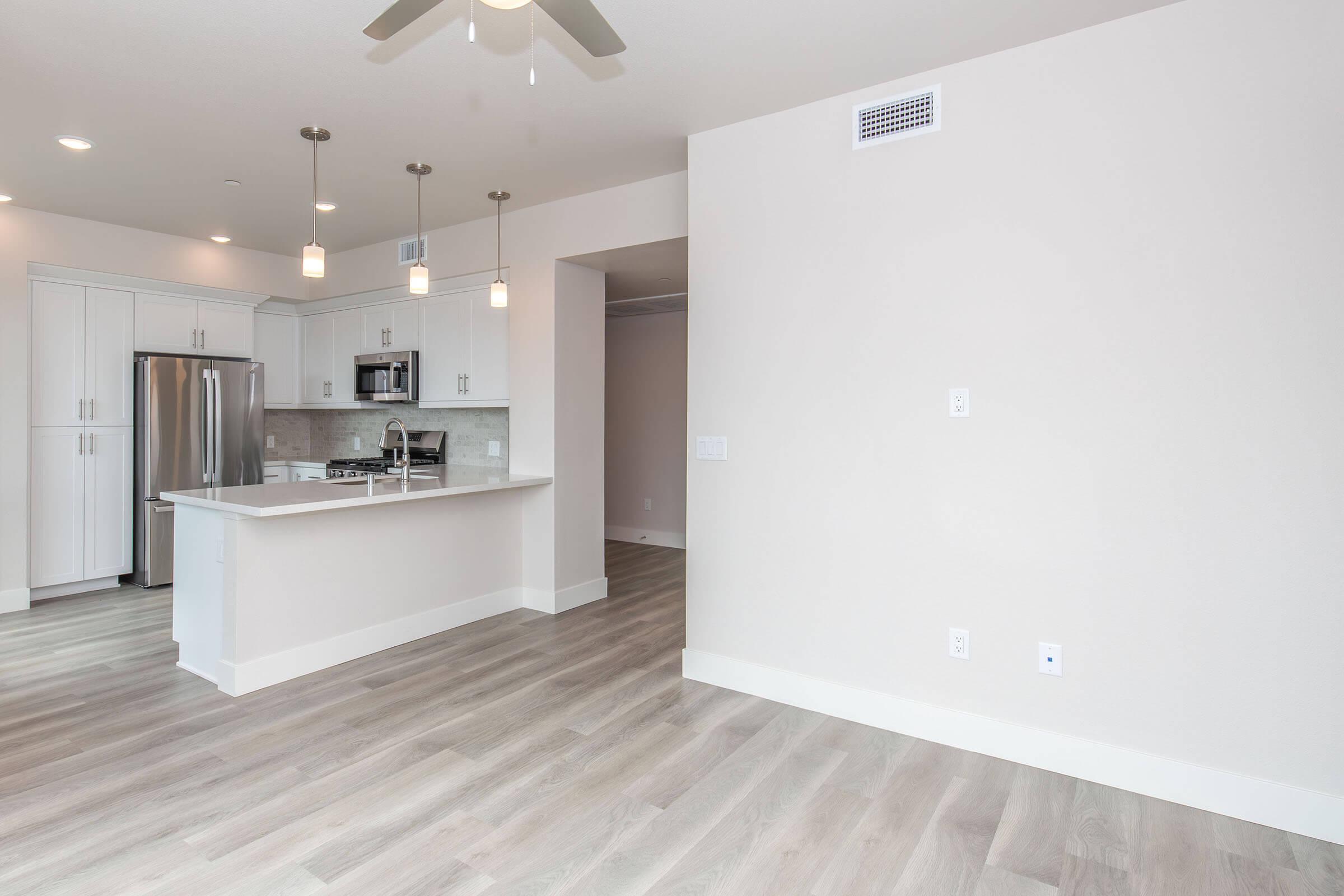 Modern kitchen and living area in an apartment. The kitchen features stainless steel appliances, a large island with pendant lighting, and light wooden flooring. The walls are painted in a neutral color, creating a bright and open atmosphere.