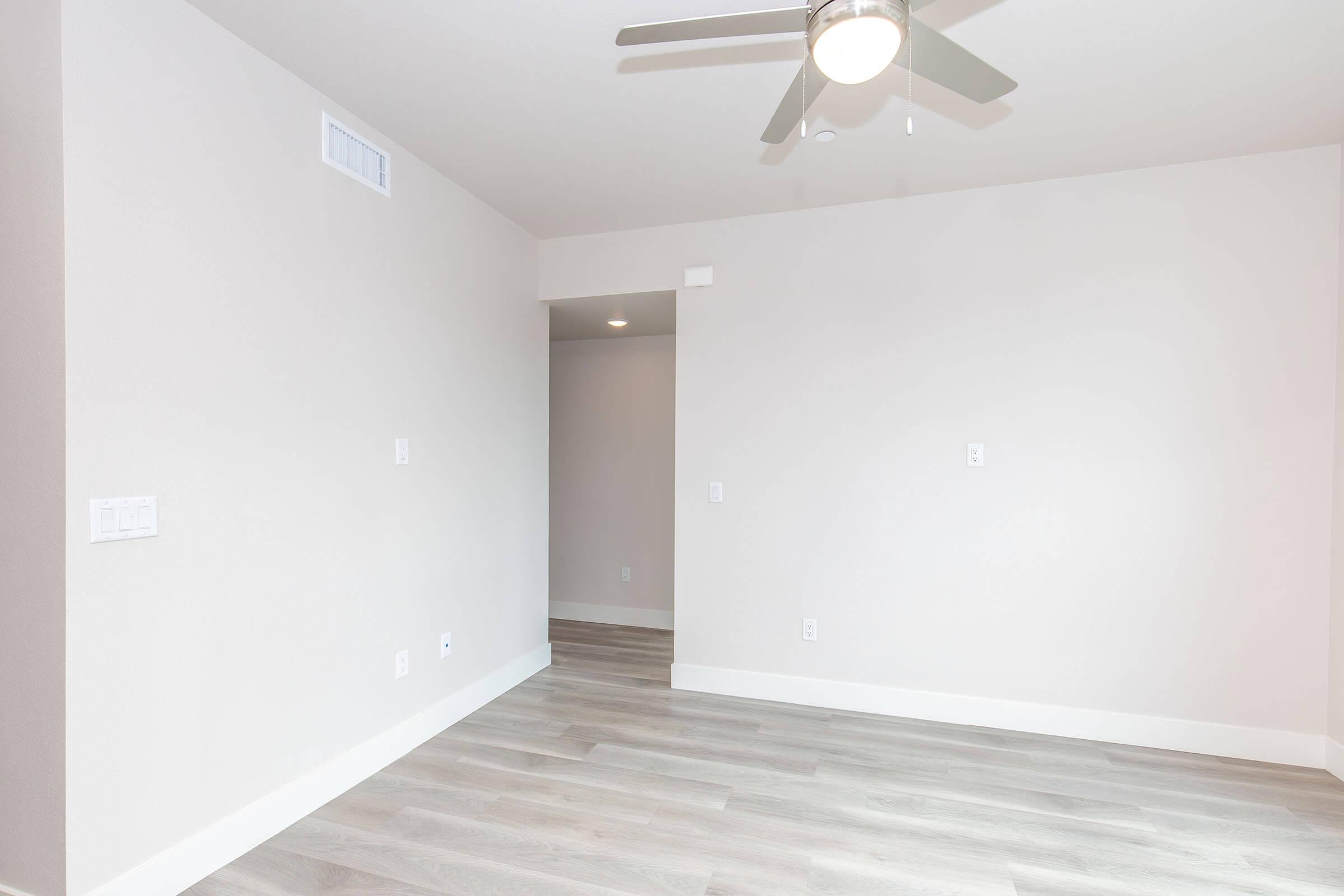 A light and airy room with a ceiling fan, featuring neutral-colored walls and light hardwood flooring. There are two wall-mounted electrical outlets and a doorway leading to another space. The overall atmosphere is clean and modern, suitable for various uses.