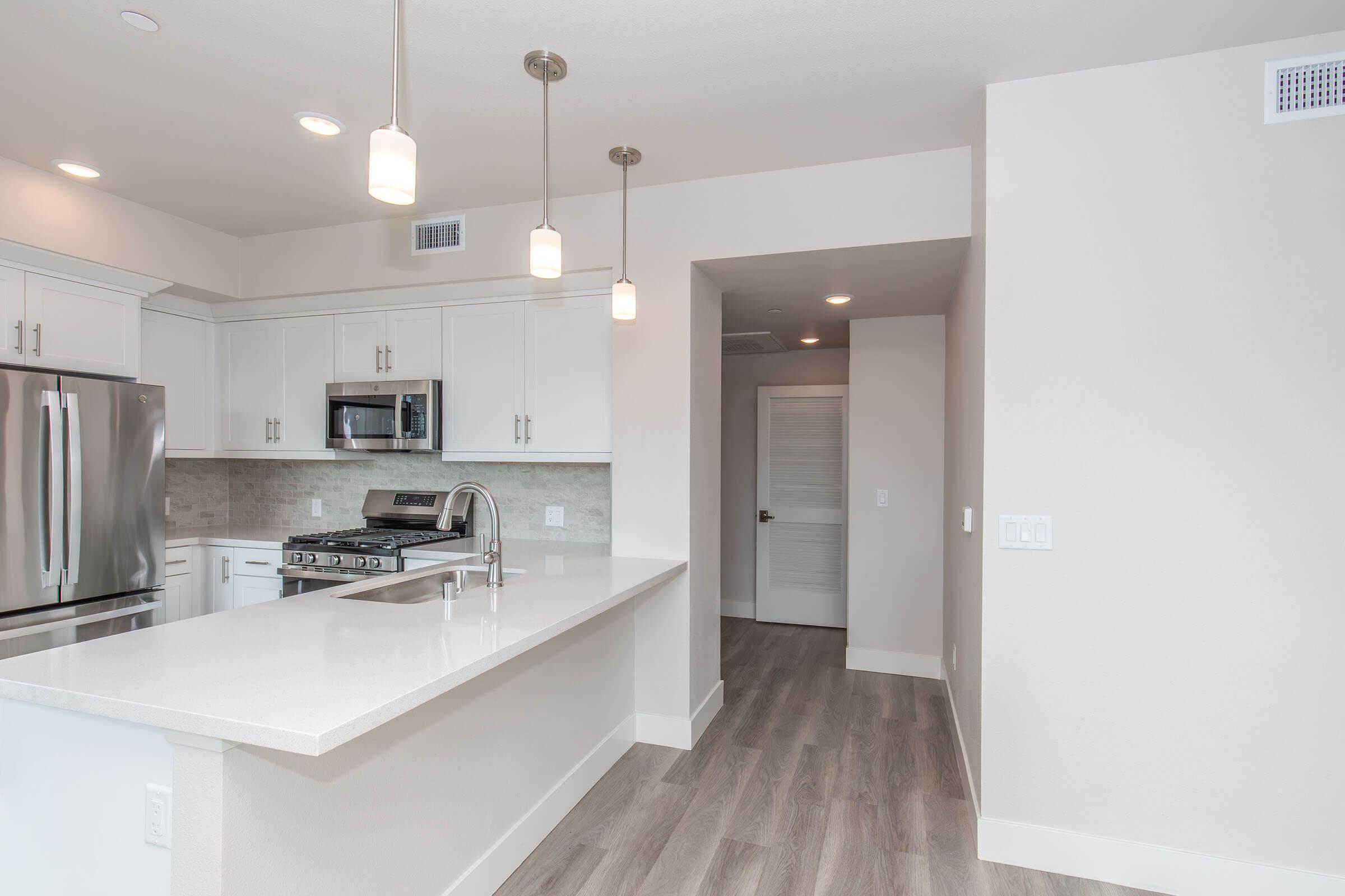 Modern kitchen featuring stainless steel appliances, white cabinetry, and a large island with a quartz countertop. Pendant lights hang above the island, and a doorway leads to an adjacent room. The space has light-colored walls and hardwood flooring for a contemporary look.