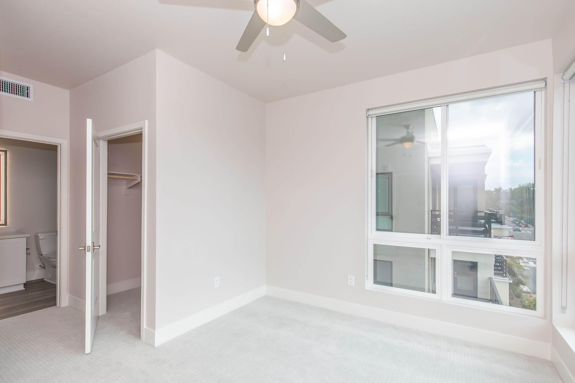 Empty interior of a contemporary bedroom featuring light-colored walls, a ceiling fan, and a large window with a view. An open door leads to a closet, and there is a glimpse of a bathroom in the background. The flooring is carpeted and well-lit by natural light.