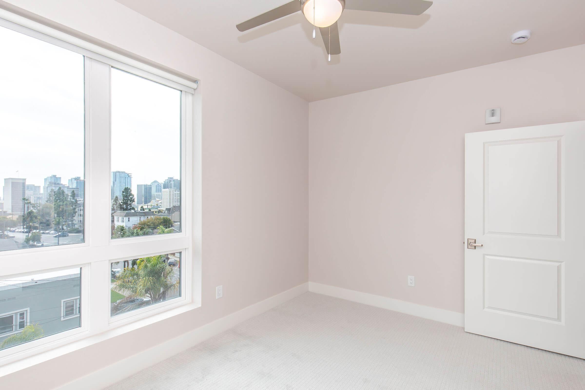 A vacant room with light beige walls and a textured carpet. It features a large window with a view of a city skyline, a ceiling fan, and a white door. Soft natural light fills the space, creating an airy atmosphere.