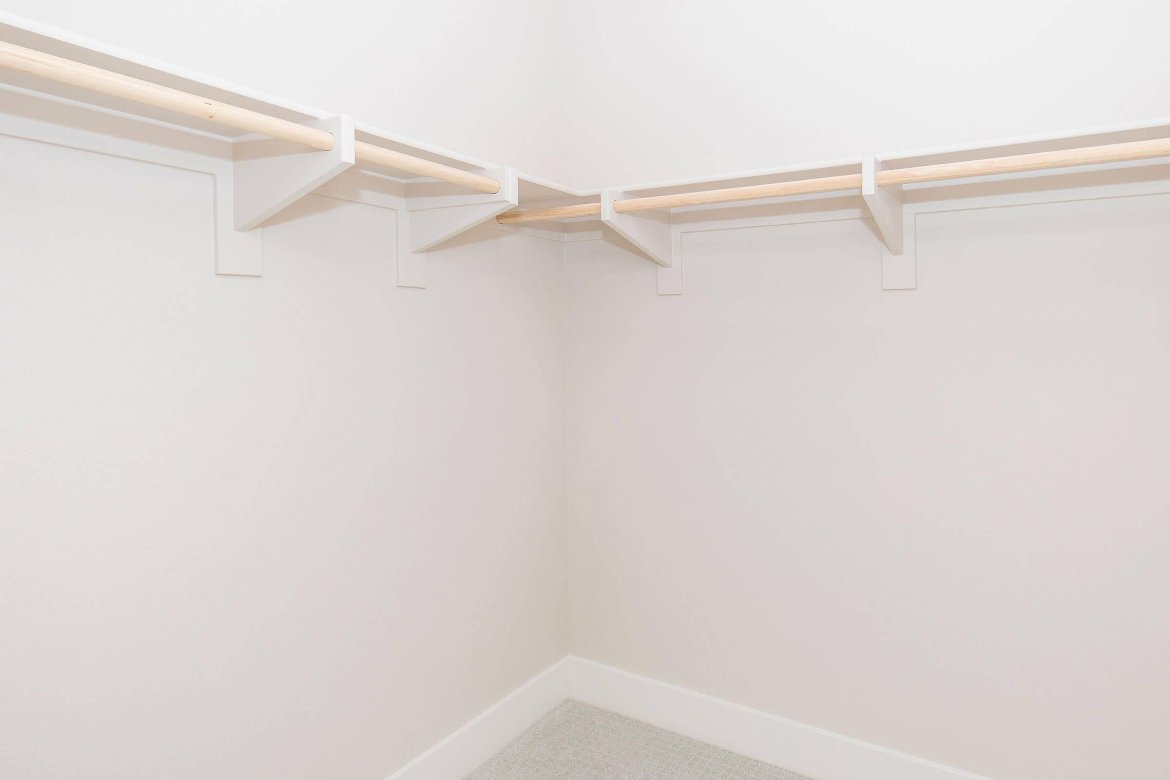 Empty closet corner with white walls and wooden rods supported by white brackets. The space has a clean, minimalistic design, featuring light-colored flooring and no visible shelves or items, creating a simple and open storage area.