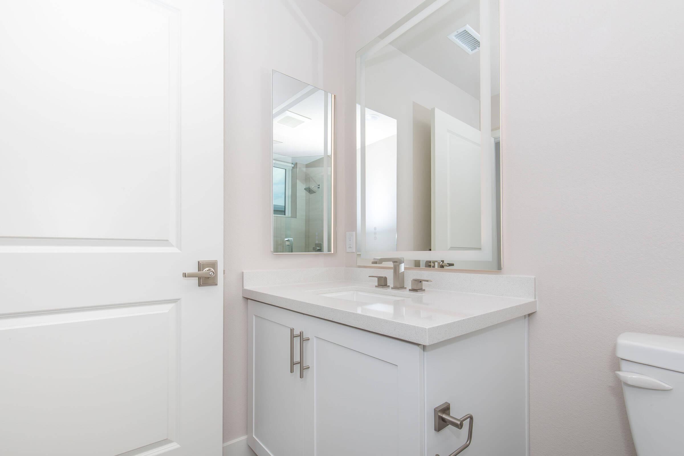A modern bathroom featuring a white vanity with dual faucets, a large mirror, and a smaller mirror mounted above. The walls are painted light and the door is partially open, showing a clean and bright space. A toilet is visible on the right side of the image.