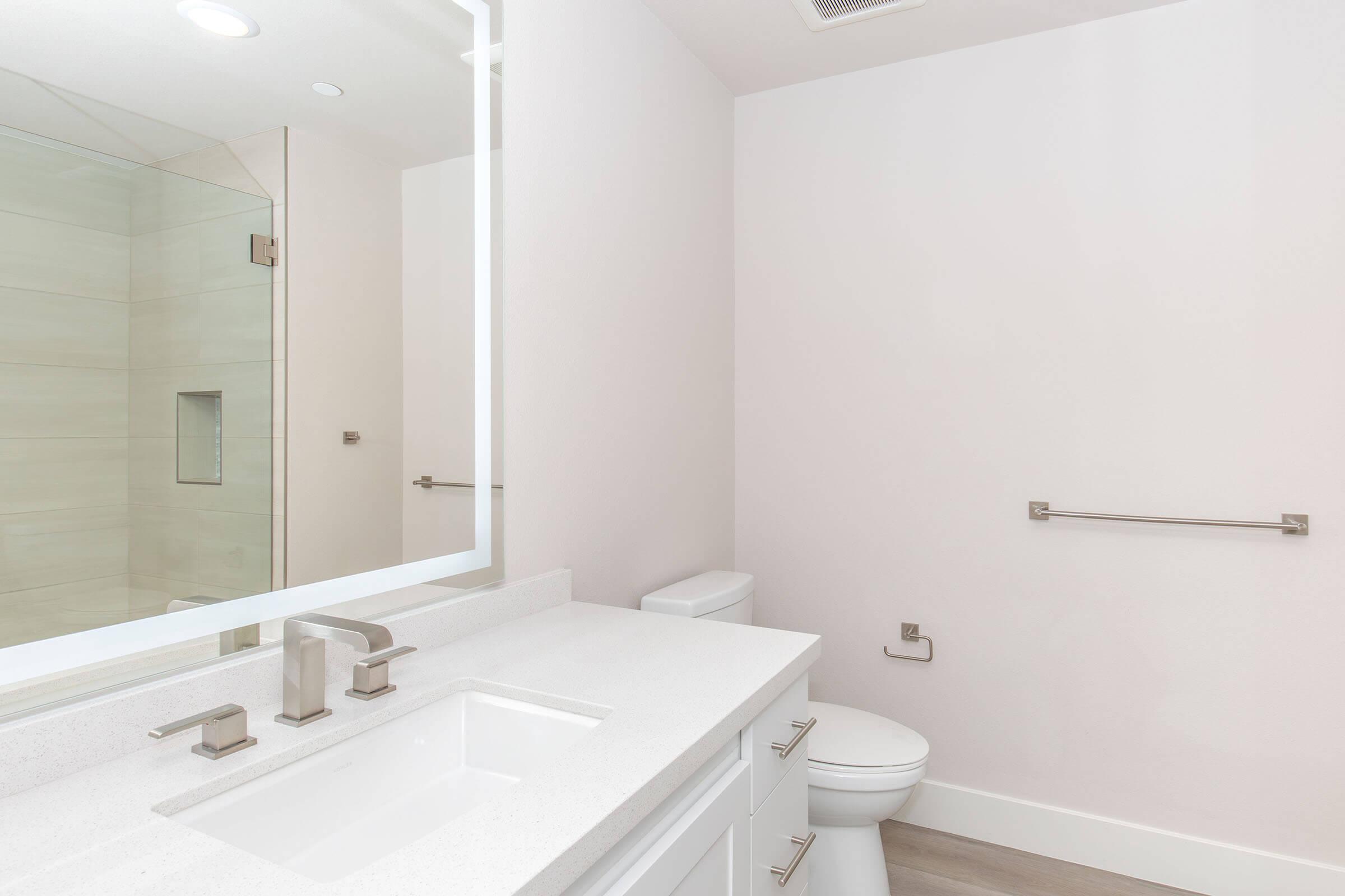 A modern bathroom featuring a white countertop with a sink and chrome faucets, a large mirror with backlighting, a toilet, and light-colored tiled walls. The decor is minimalist, with neutral tones and contemporary fixtures. Natural light may be coming from a window or overhead lighting.