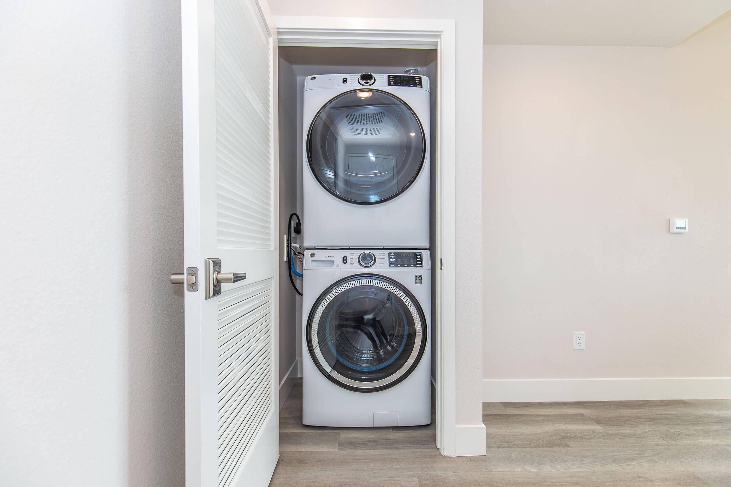 A stacked washer and dryer set located in a spacious laundry closet with a white door. The appliances have a modern design, featuring large front-loading doors and a clean, minimalist aesthetic. Light-colored walls and wood flooring enhance the overall brightness of the space.