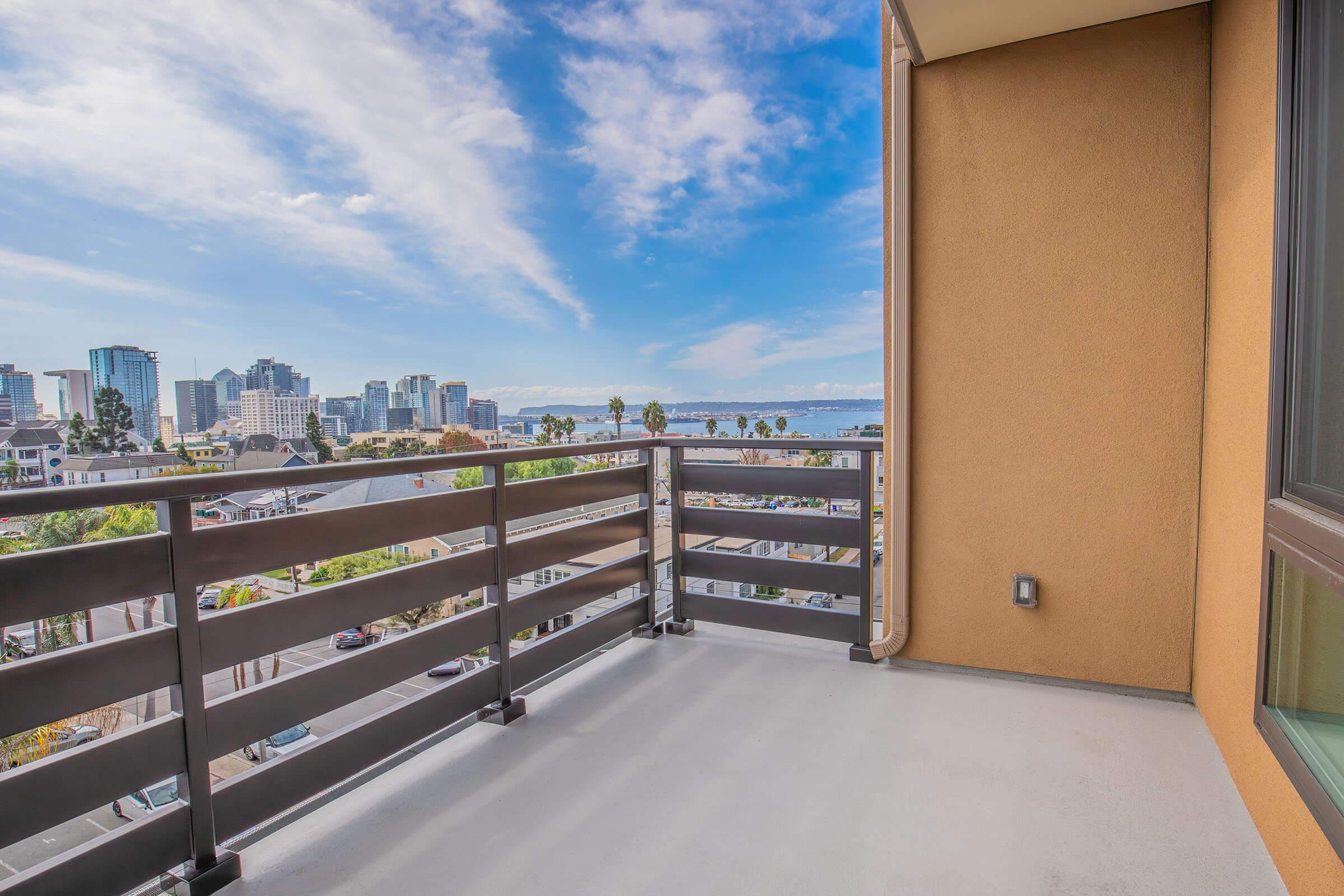 View from a balcony overlooking a city skyline with tall buildings, palm trees, and a body of water in the distance, under a blue sky with clouds. The balcony features a modern railing design against a beige wall.