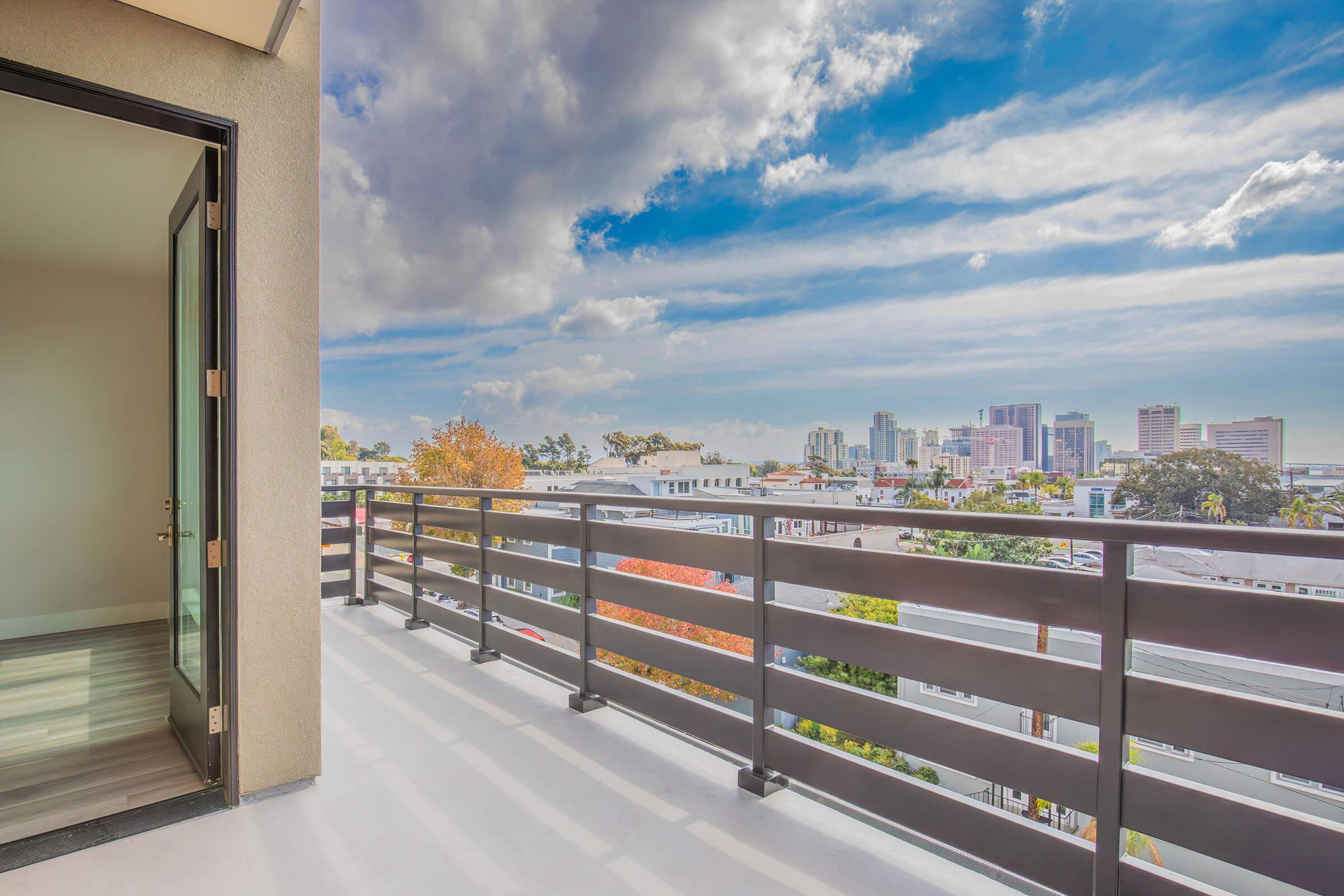 A spacious balcony with a modern railing, overlooking a vibrant city skyline. The scene features a bright blue sky with scattered clouds and hints of colorful trees below. The open door suggests an inviting indoor space just inside.