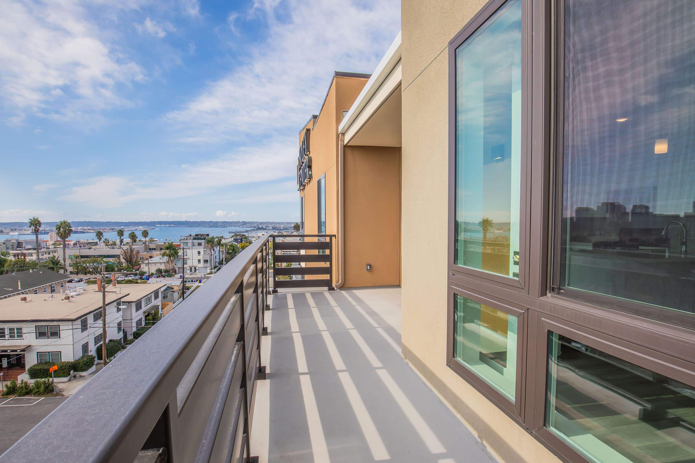 A spacious balcony with a railing overlooking a coastal view. The scene features a bright blue sky with scattered clouds, and the ocean is visible in the distance. Nearby buildings and palm trees are seen below, adding to the lively atmosphere of the area.