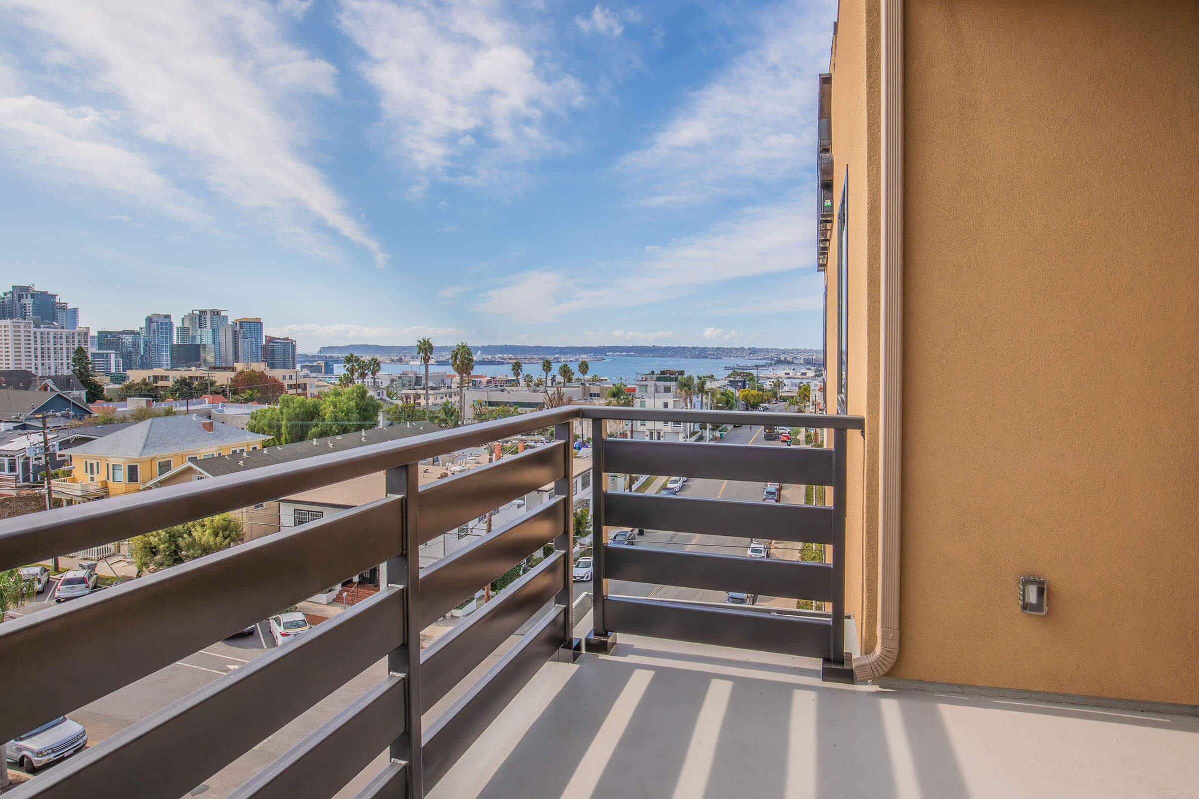 View from a balcony overlooking a coastal cityscape with modern buildings, palm trees, and a marina. The sky is clear with some clouds, showcasing a sunny day. The balcony features a metal railing, emphasizing the outdoor space.