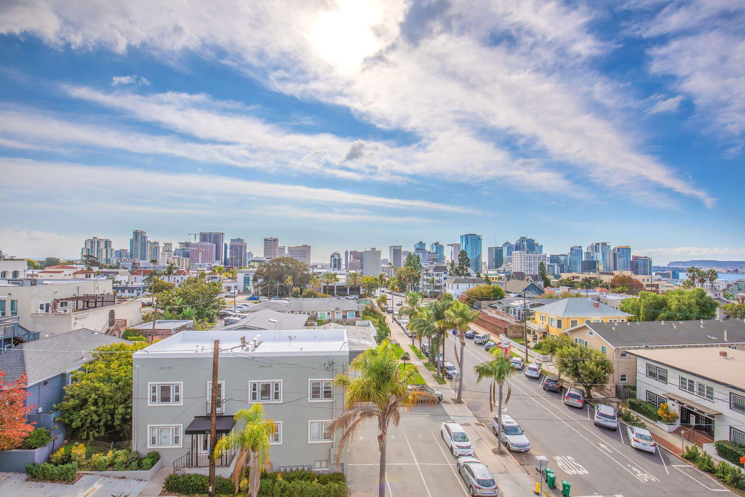 A vibrant city skyline with modern skyscrapers under a blue sky with wispy clouds. In the foreground, palm trees line a residential street with a mix of houses and parked cars. The scene captures a lively urban atmosphere, showcasing a blend of nature and city life.