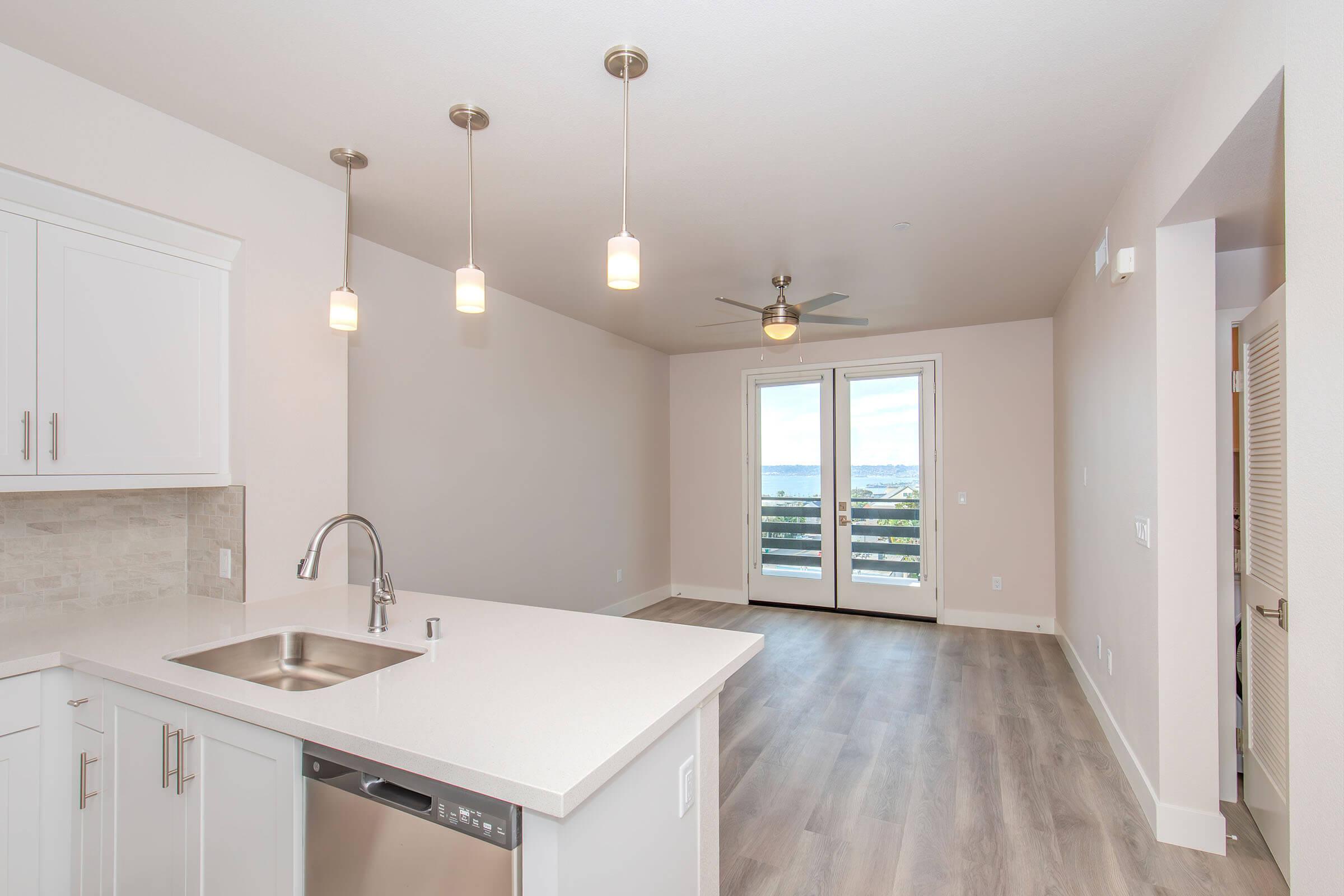 A modern kitchen with a sleek countertop and stainless steel appliances, featuring pendant lighting above. The open layout leads to a living area with a ceiling fan and large glass doors opening to a balcony, offering a view of the outdoors. The decor is light and contemporary, highlighted by warm wood flooring.