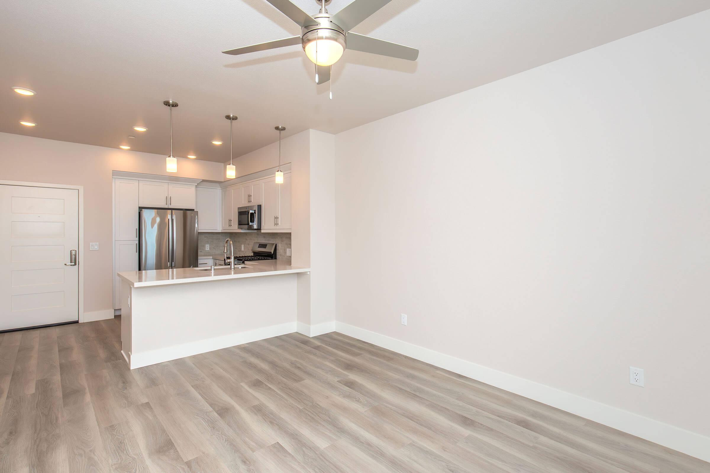 A modern kitchen and living area featuring a light-colored open space, hardwood-like flooring, stainless steel appliances, white cabinetry, and pendant lighting. The kitchen island has seating space, and the room is bright with natural light from the ceiling fixtures.
