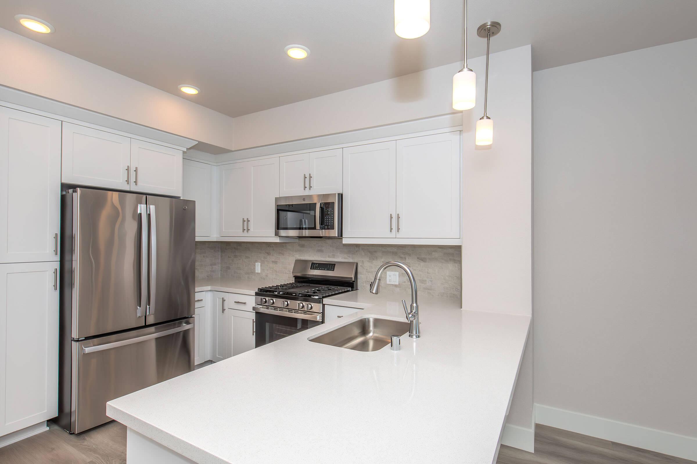 Modern kitchen featuring stainless steel appliances, including a refrigerator, microwave, and gas stove. The white cabinetry contrasts with a light gray backsplash. A countertop island provides additional workspace, and pendant lights illuminate the area, creating a bright and inviting atmosphere.