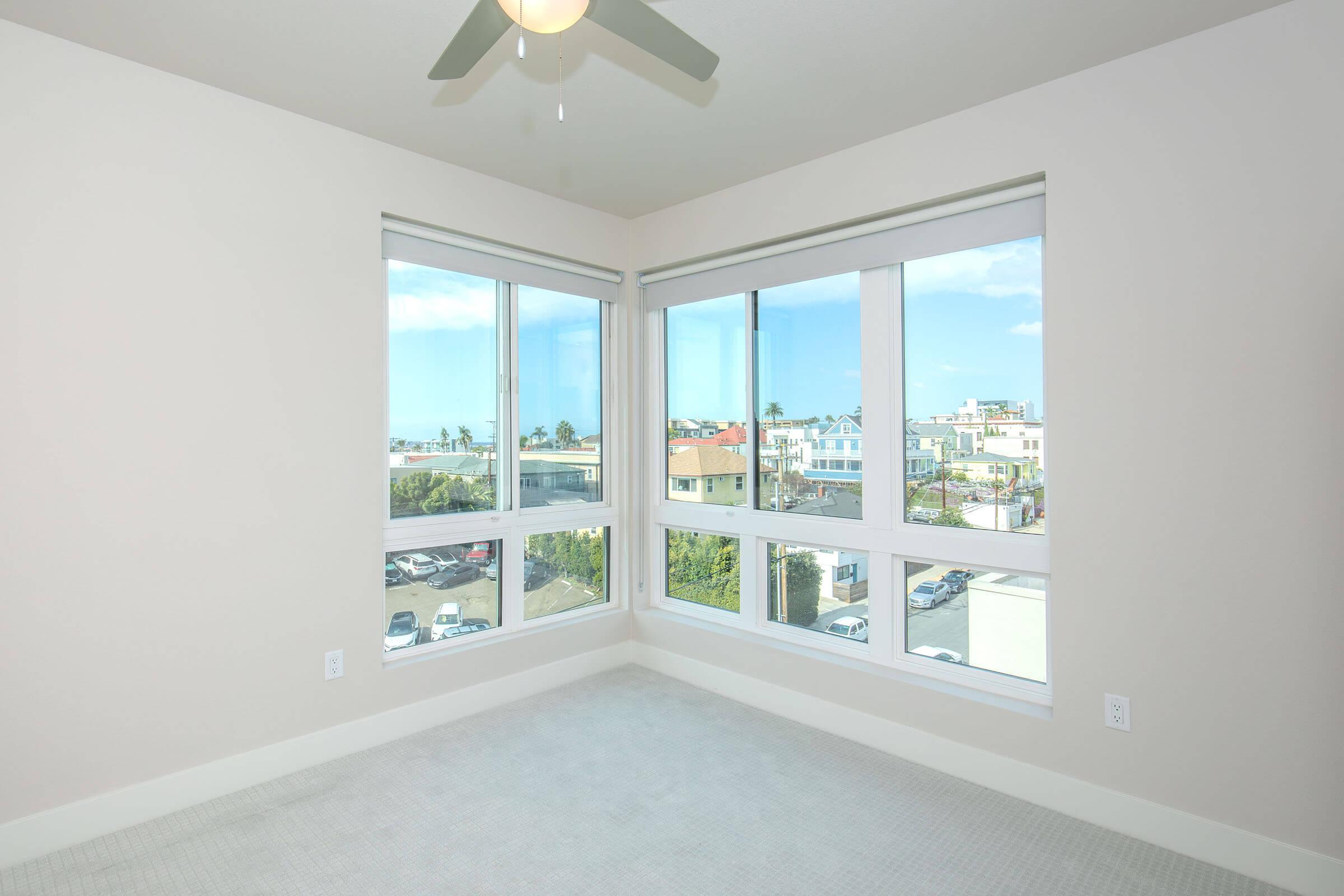A bright, empty bedroom with large windows showcasing a view of houses and greenery outside. The room features a light-colored carpet and a ceiling fan, creating a spacious and airy atmosphere. Natural light floods the space, highlighting the clean walls and modern windows.
