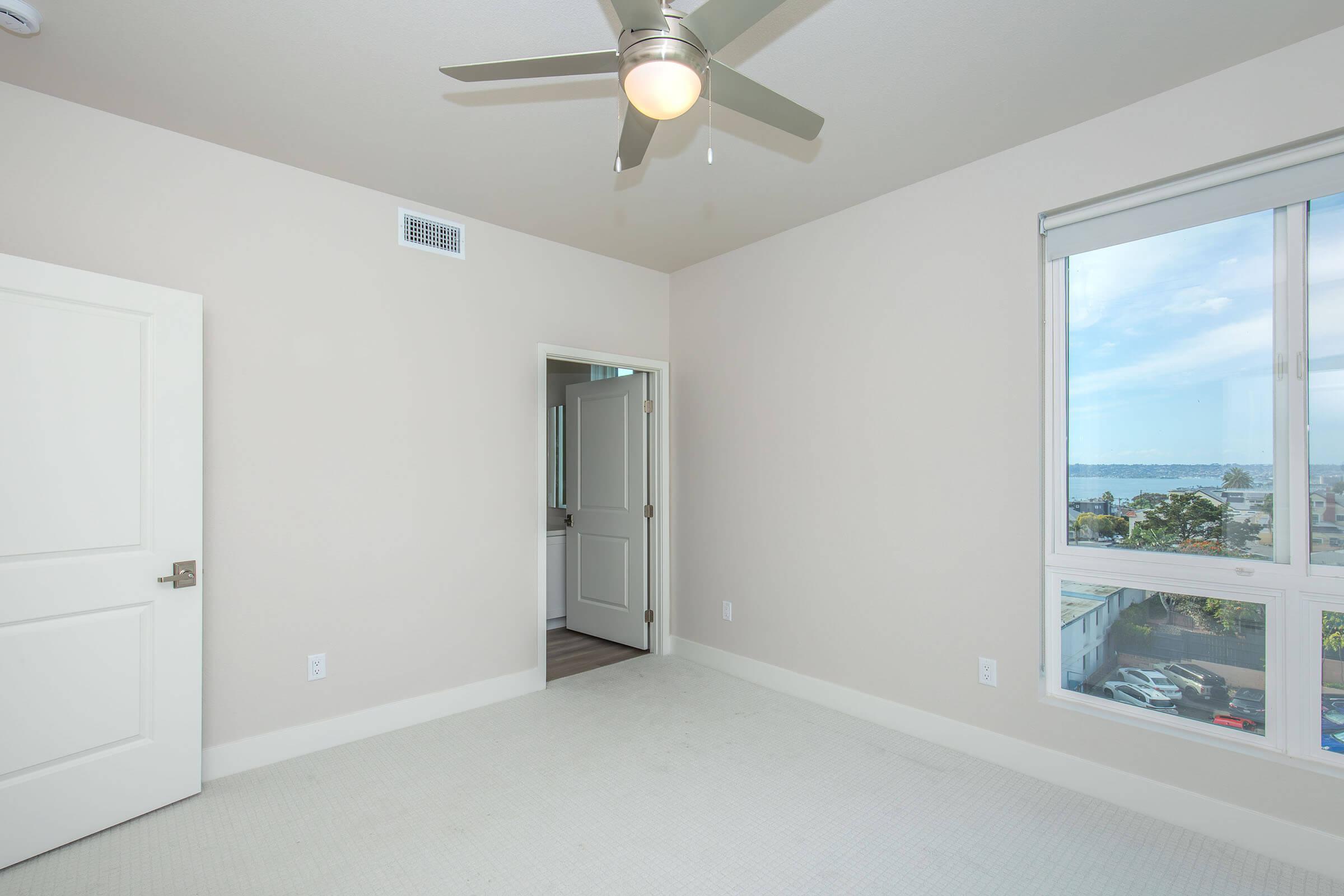 Empty room with light-colored walls and carpet, featuring a ceiling fan and a window with a view of the outside. A door on the left leads to another room, and natural light fills the space, creating an airy atmosphere.